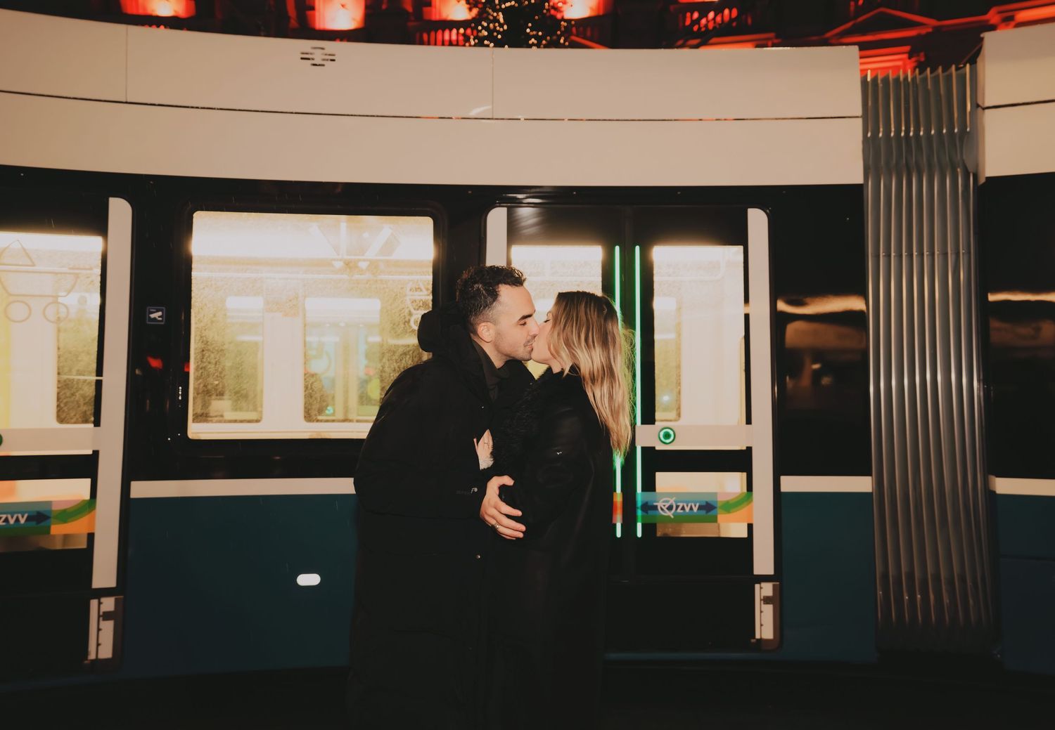 Couple sharing a tender kiss inside a warmly lit metro station with a tram visible behind them.