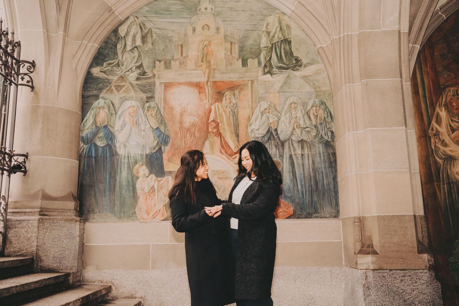 Mom and daughter standing together beneath an arched doorway with elaborate religious fresco artwork.