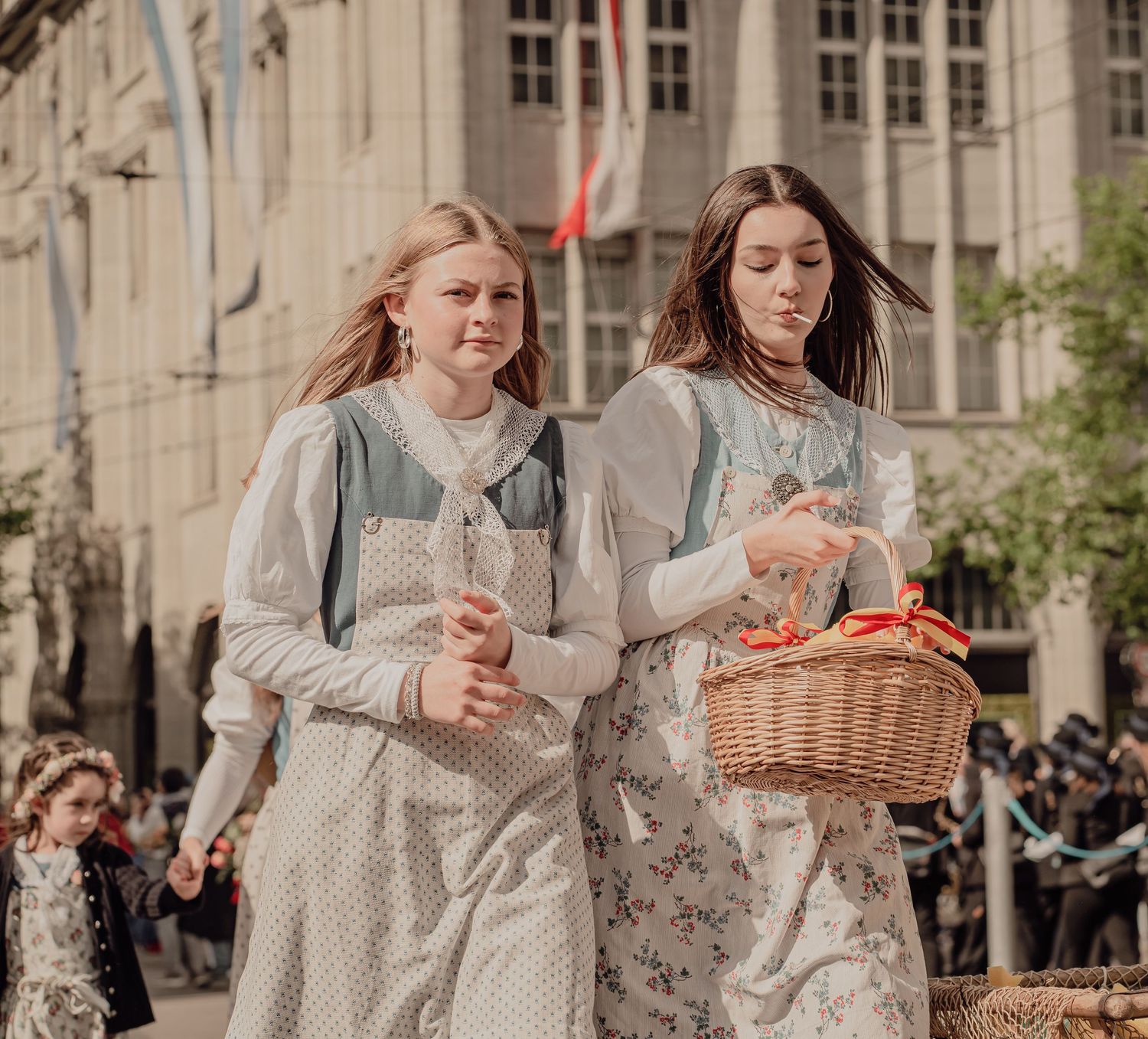 Two young women in vintage floral dresses walk together carrying a wicker basket in a historical parade.