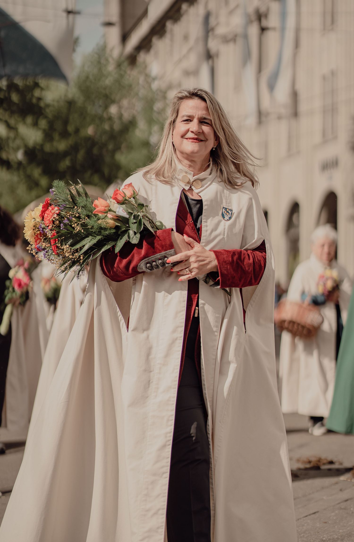 Woman in white ceremonial robe smiles holding colorful bouquet during outdoor festival parade.