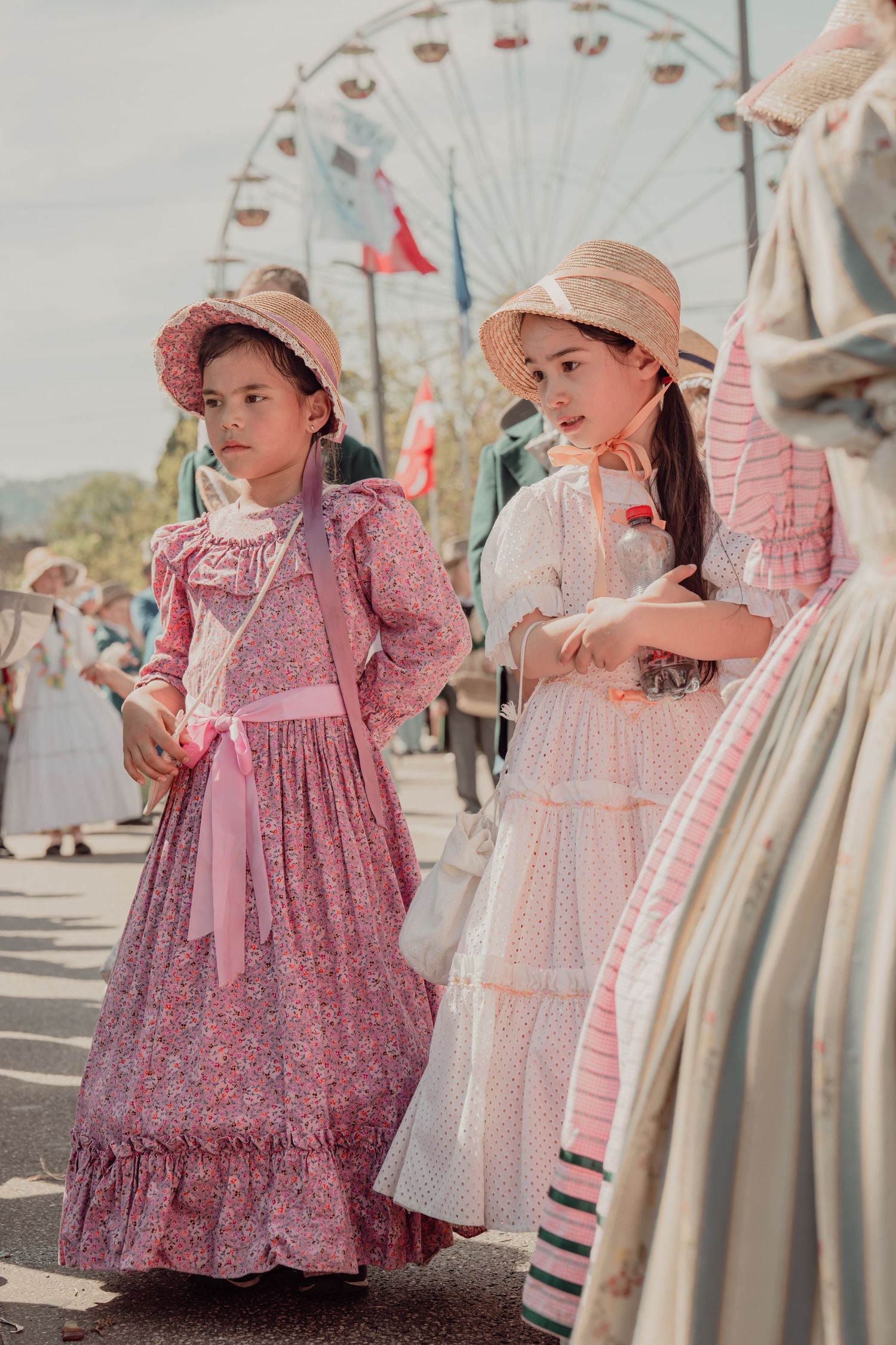 Two girls in vintage pink floral dresses and straw bonnets walk near a ferris wheel at festival.
