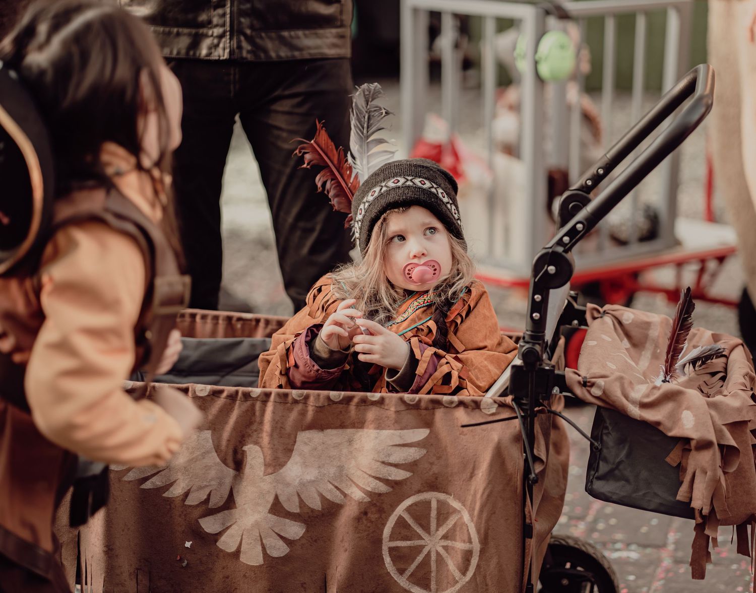 Toddler in Native American costume with pacifier rides in a decorated stroller at carnival.