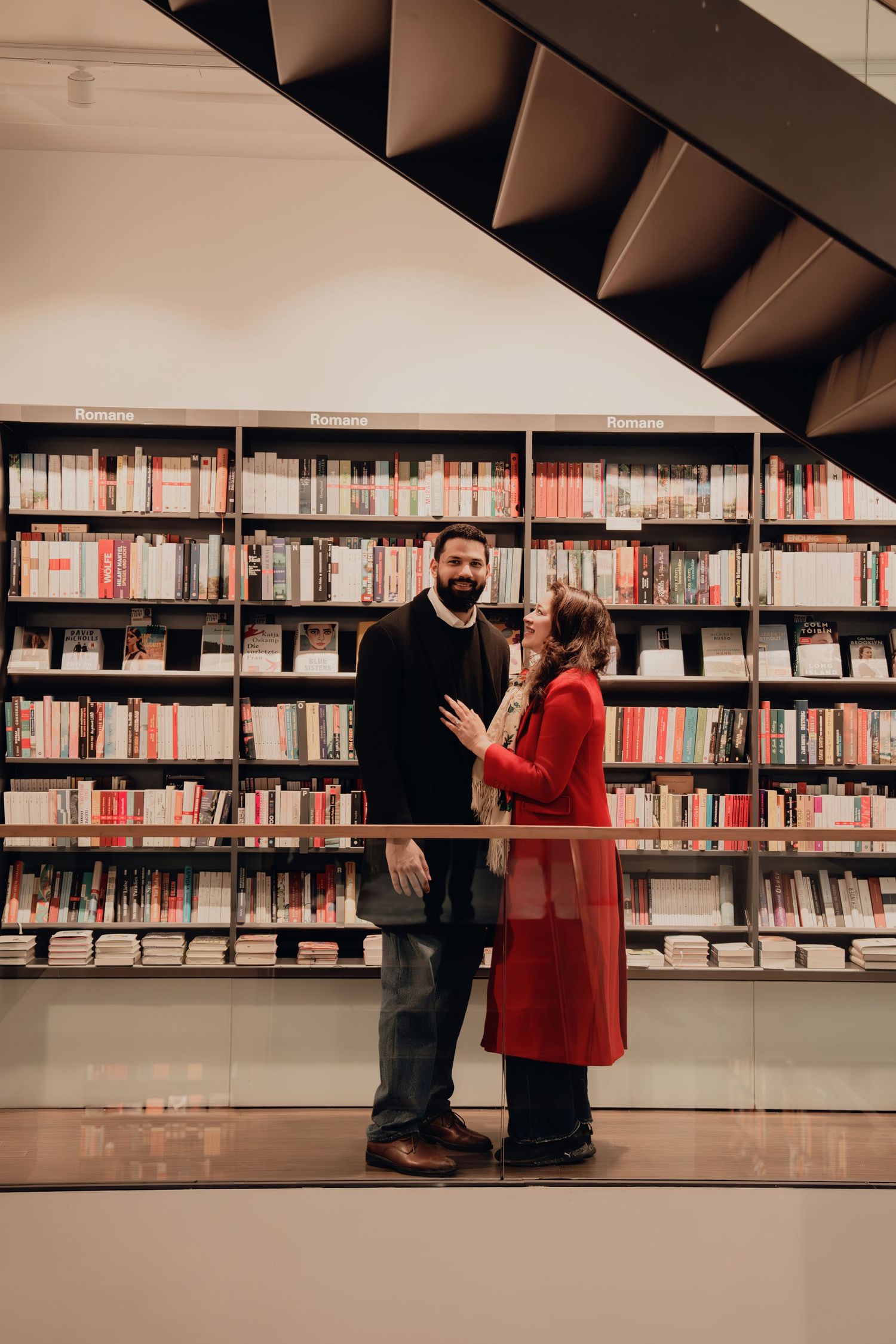 Couple smiling together in modern library with colorful bookshelves, man in black and woman in red coat.