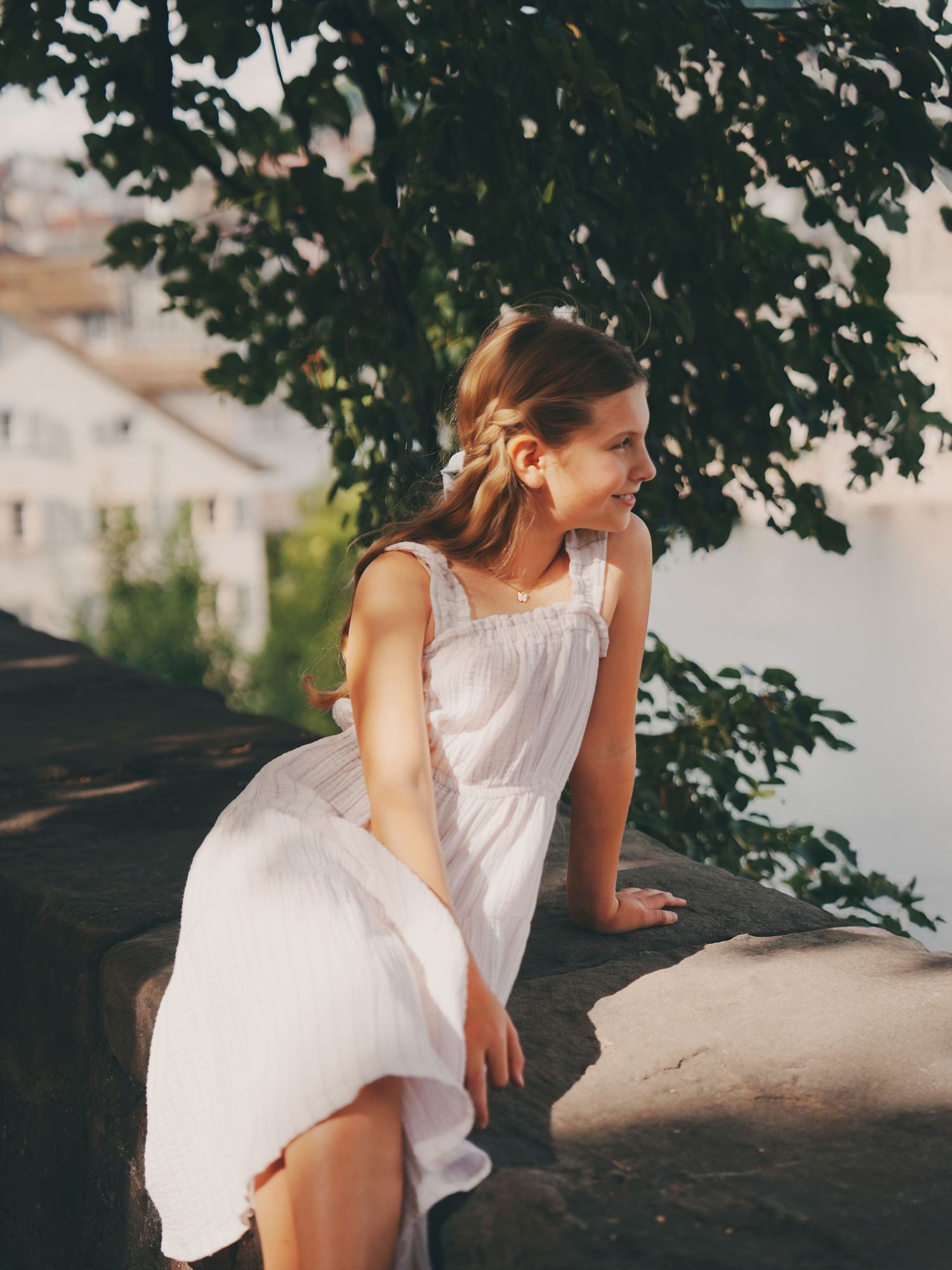 A young girl in a white dress sits on a stone wall smiling, with a cityscape behind her.