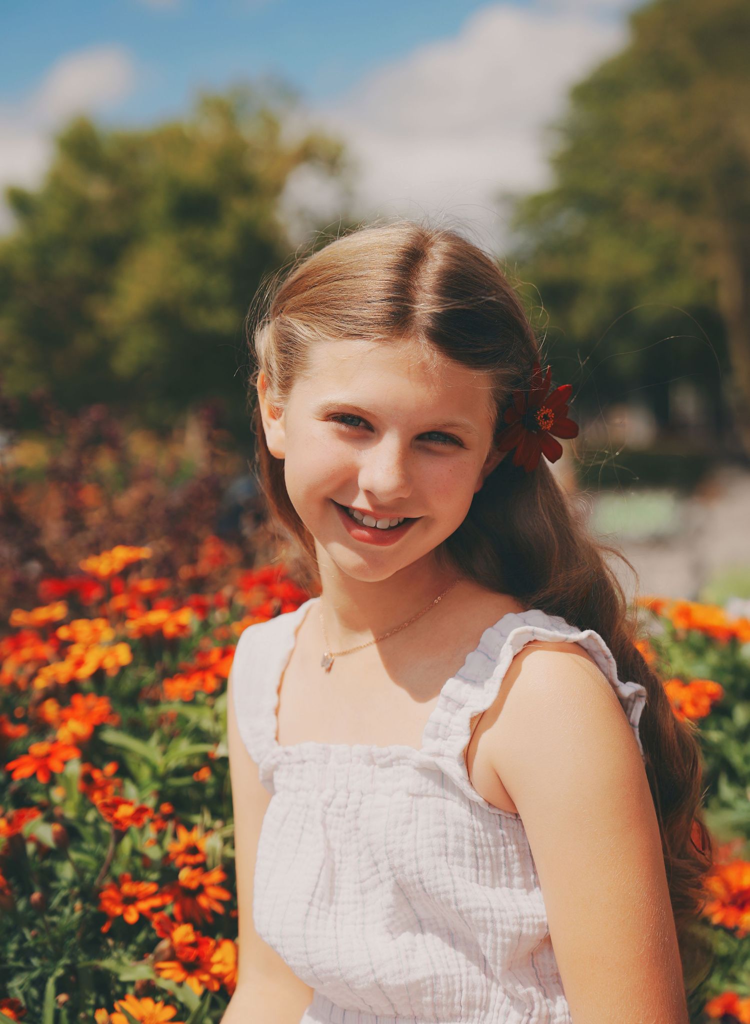Smiling young girl in white dress posing before vibrant orange and red garden flowers.