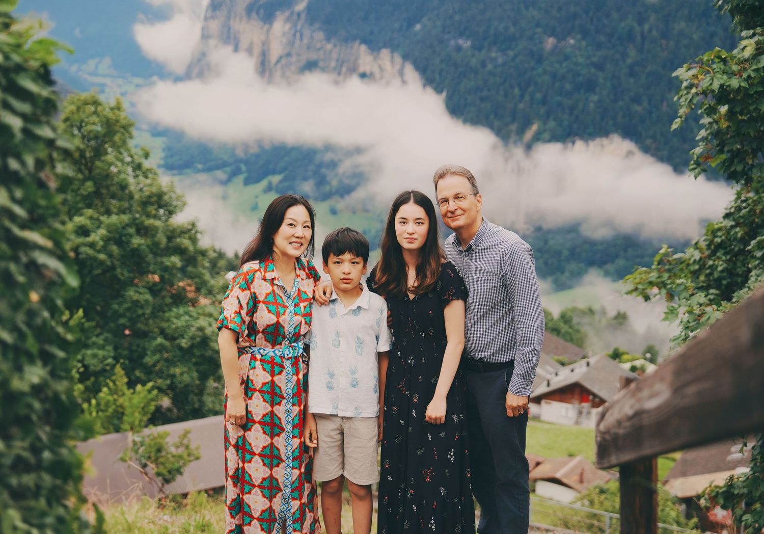 Two families pose together outdoors with lush green Swiss Alps and dramatic cloudy peaks behind them.