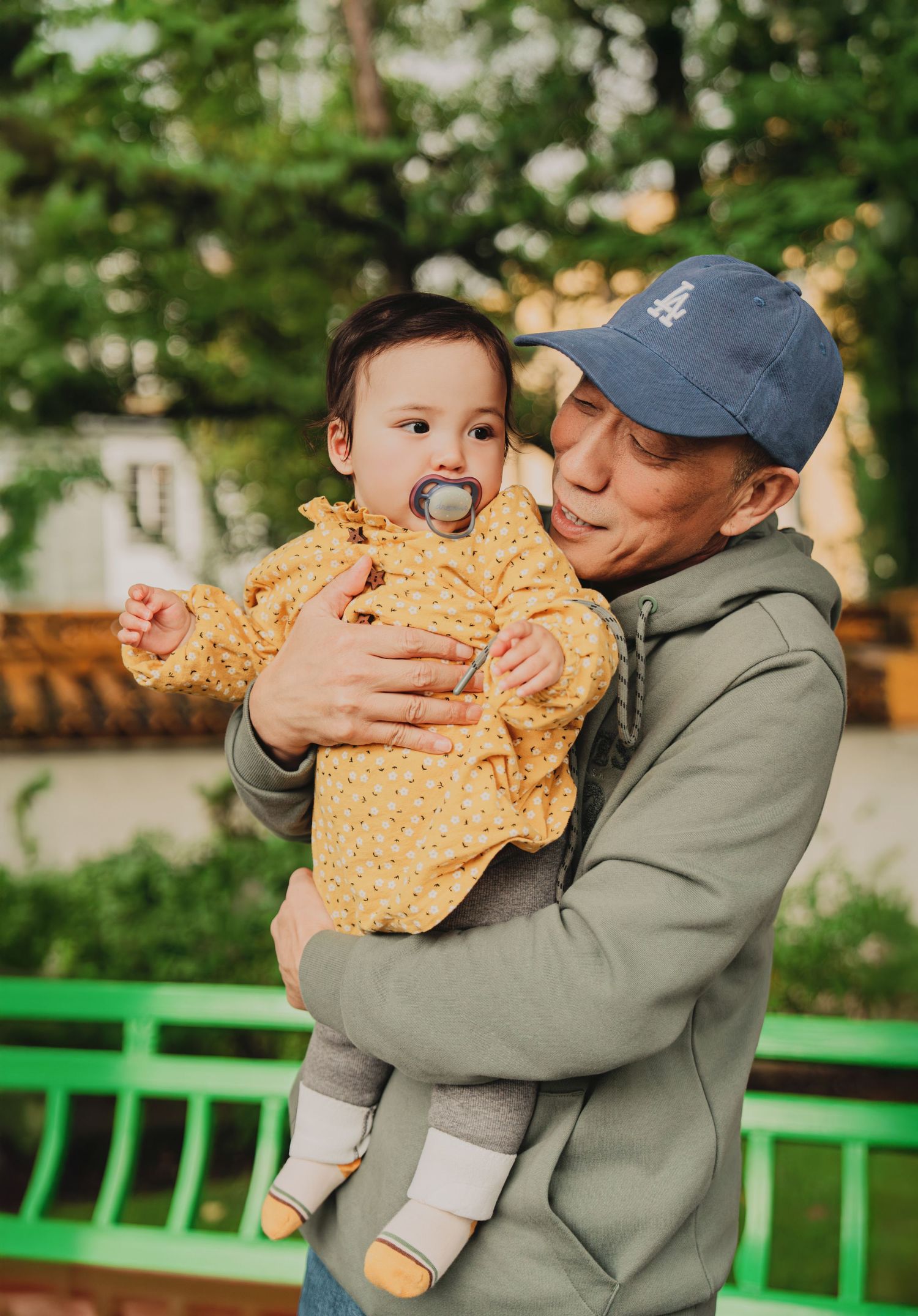 Father in blue cap tenderly holds baby with pacifier outdoors in a green park setting.