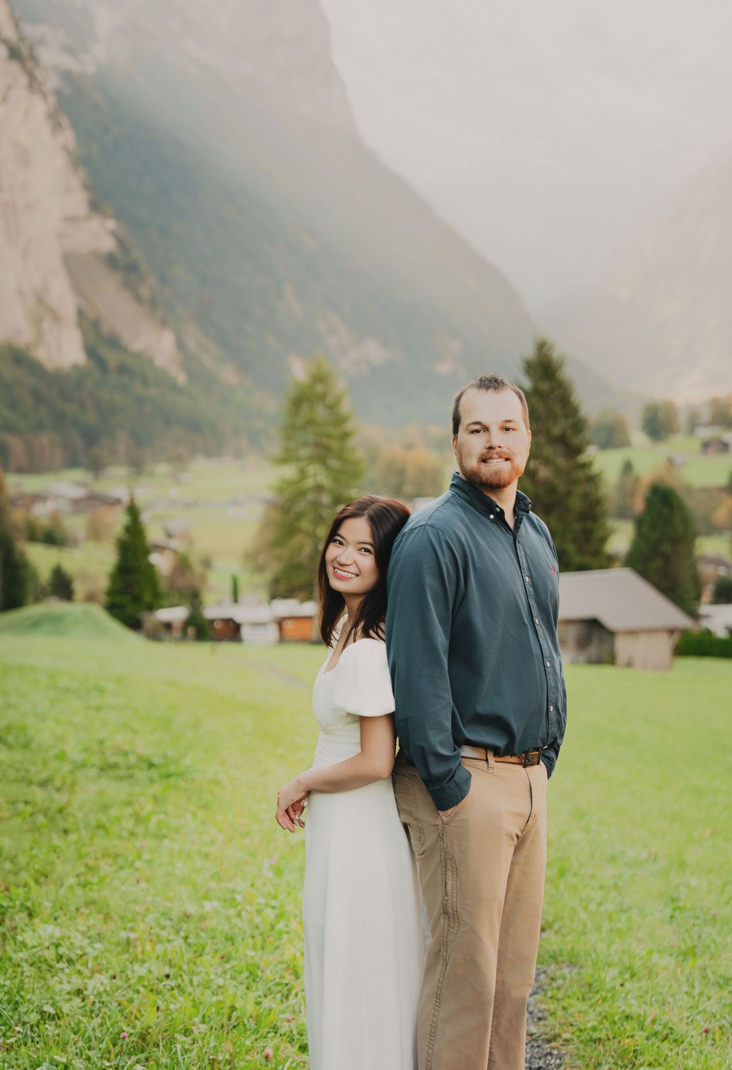 Romantic couple smiling together on a grassy field with a charming Swiss mountain village and misty peaks behind them.