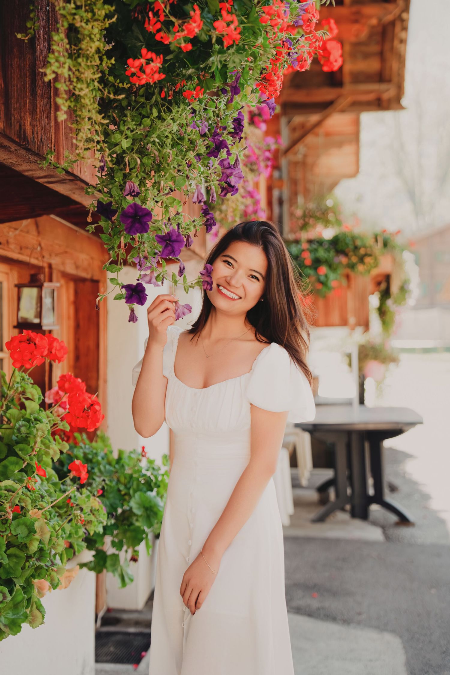 Cheerful young woman in a white dress posing beside lush hanging flower baskets with red and purple blooms outdoors.