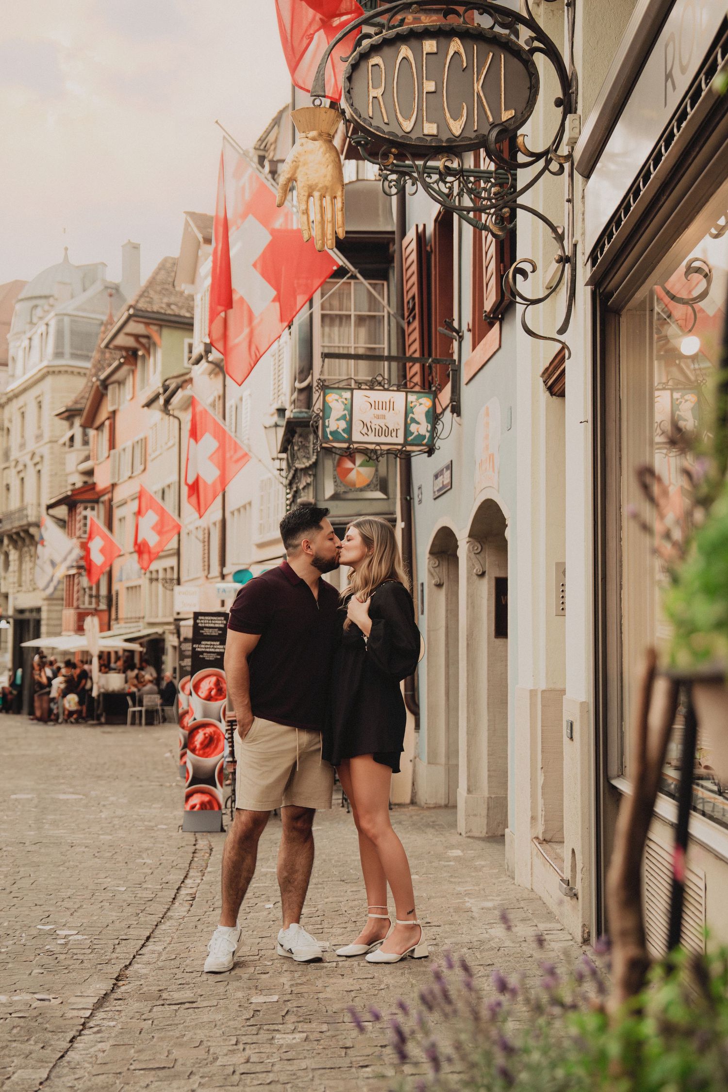 Romantic couple kissing on cobblestone street beside ROECKL shop with Swiss flags above.