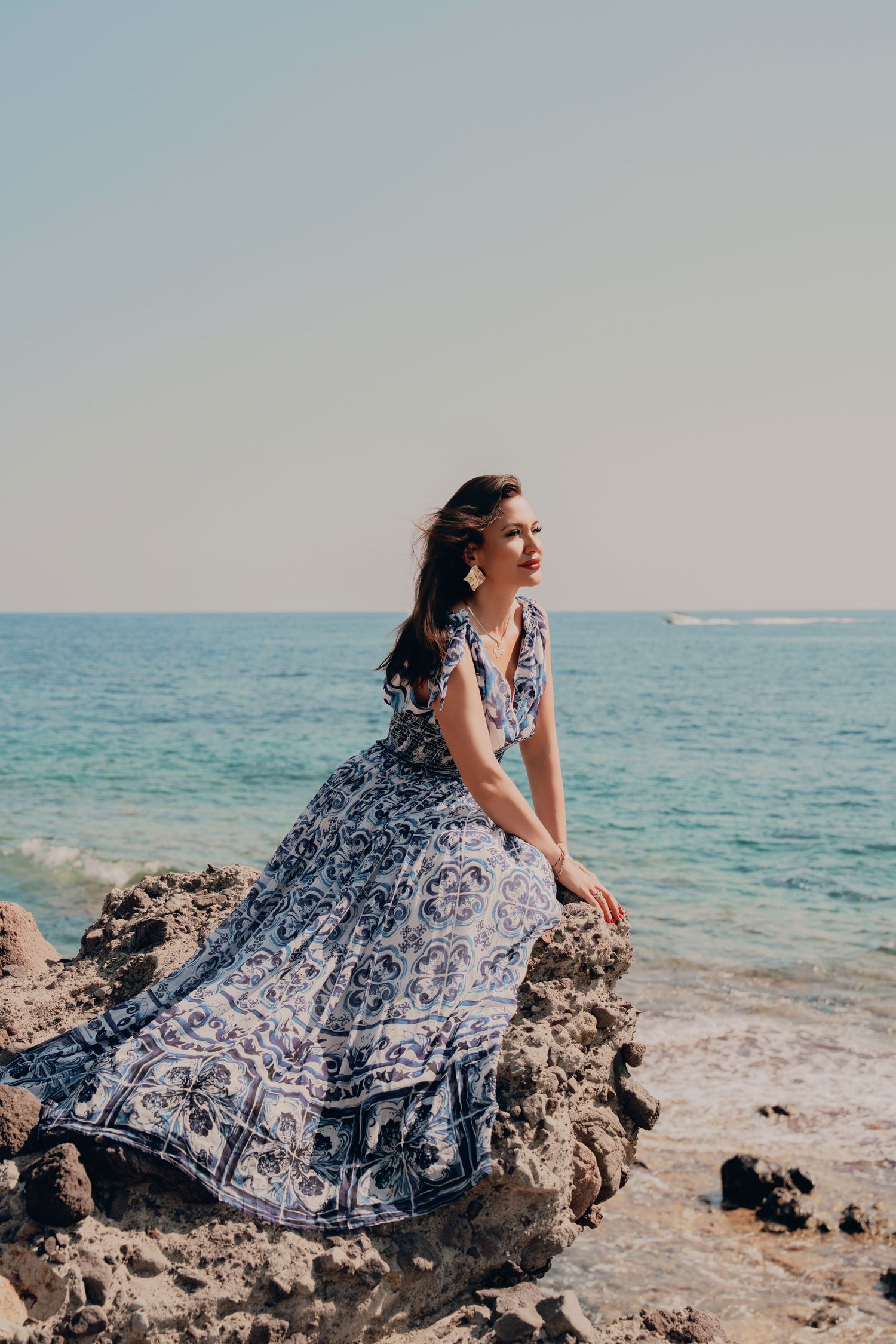 Woman in blue and white floral maxi dress sitting on coastal rocks overlooking calm ocean horizon.