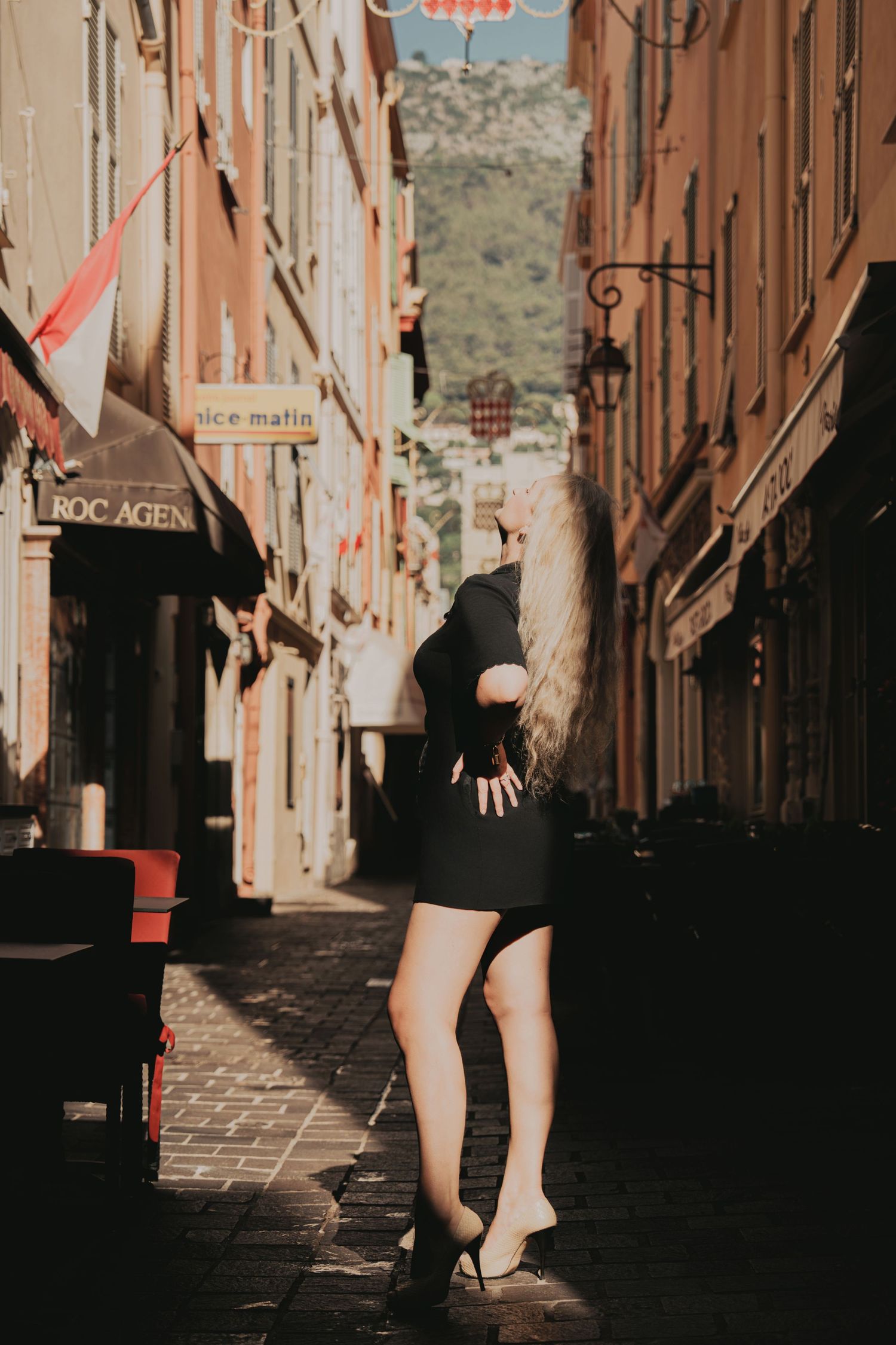 Blonde woman in black dress and heels walking away in a sunny European cobblestone alley.