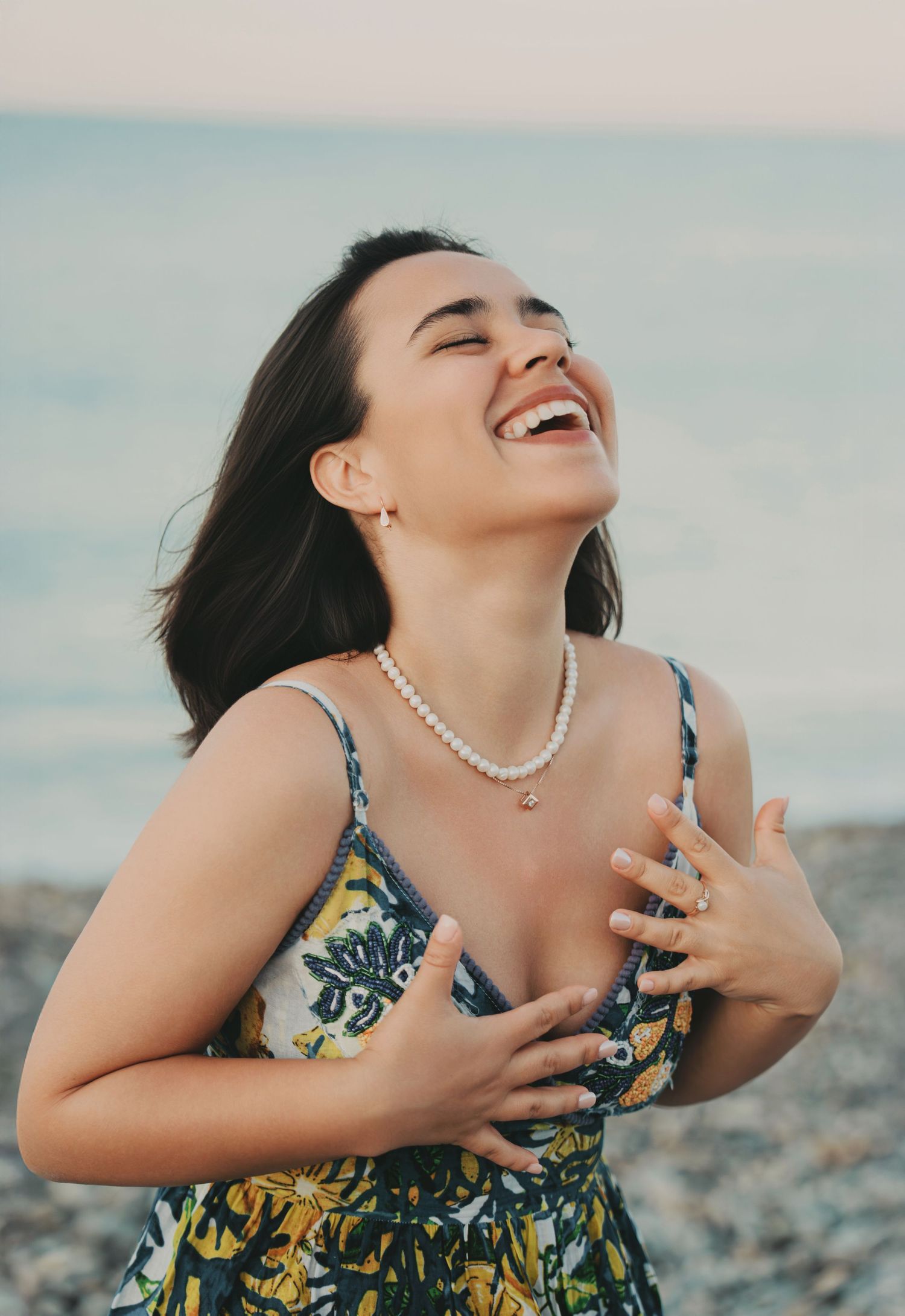A joyful woman in a floral dress laughs with a pearl necklace on a beach.