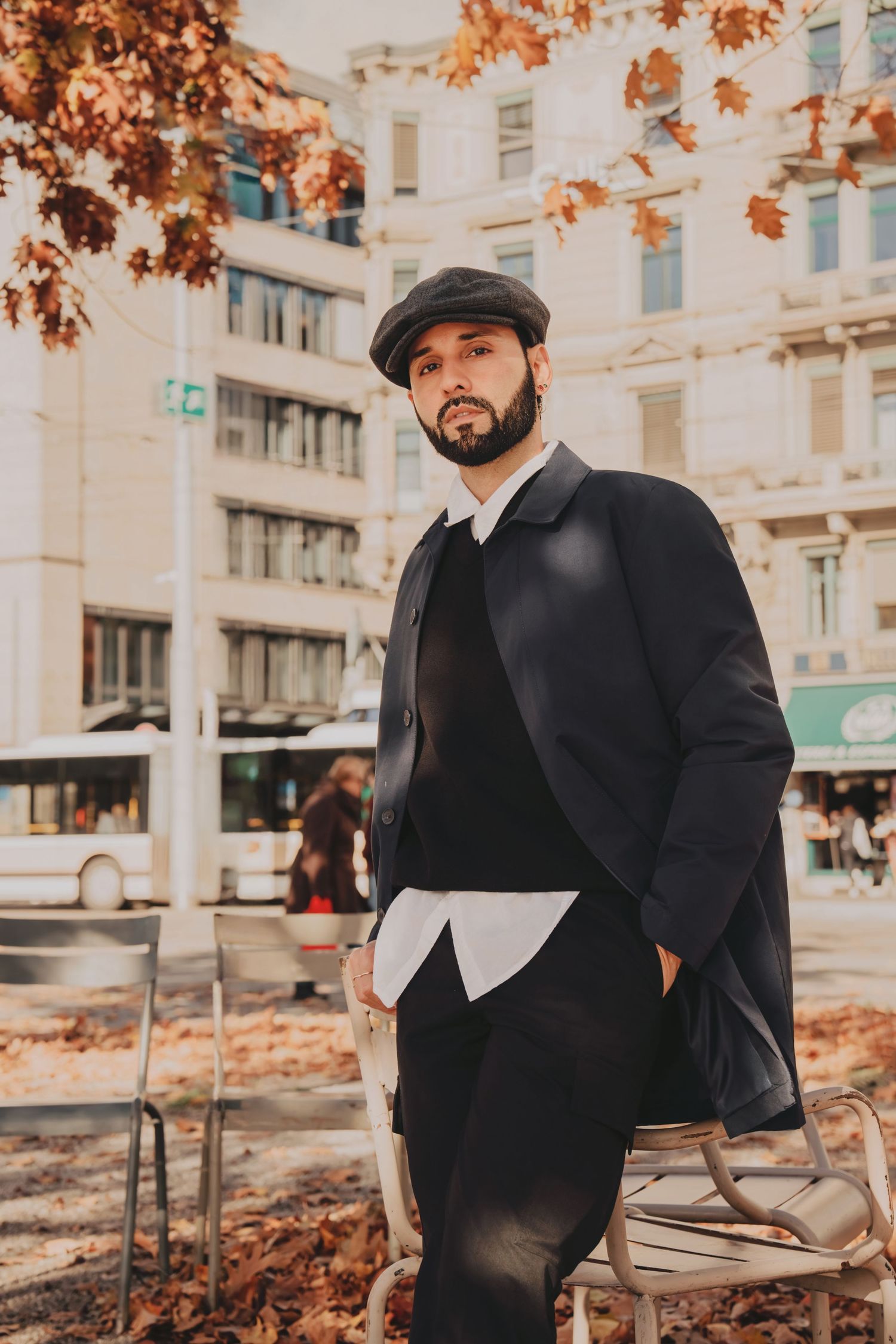 Bearded man in dark coat and newsboy cap leaning on chair in autumn city square.