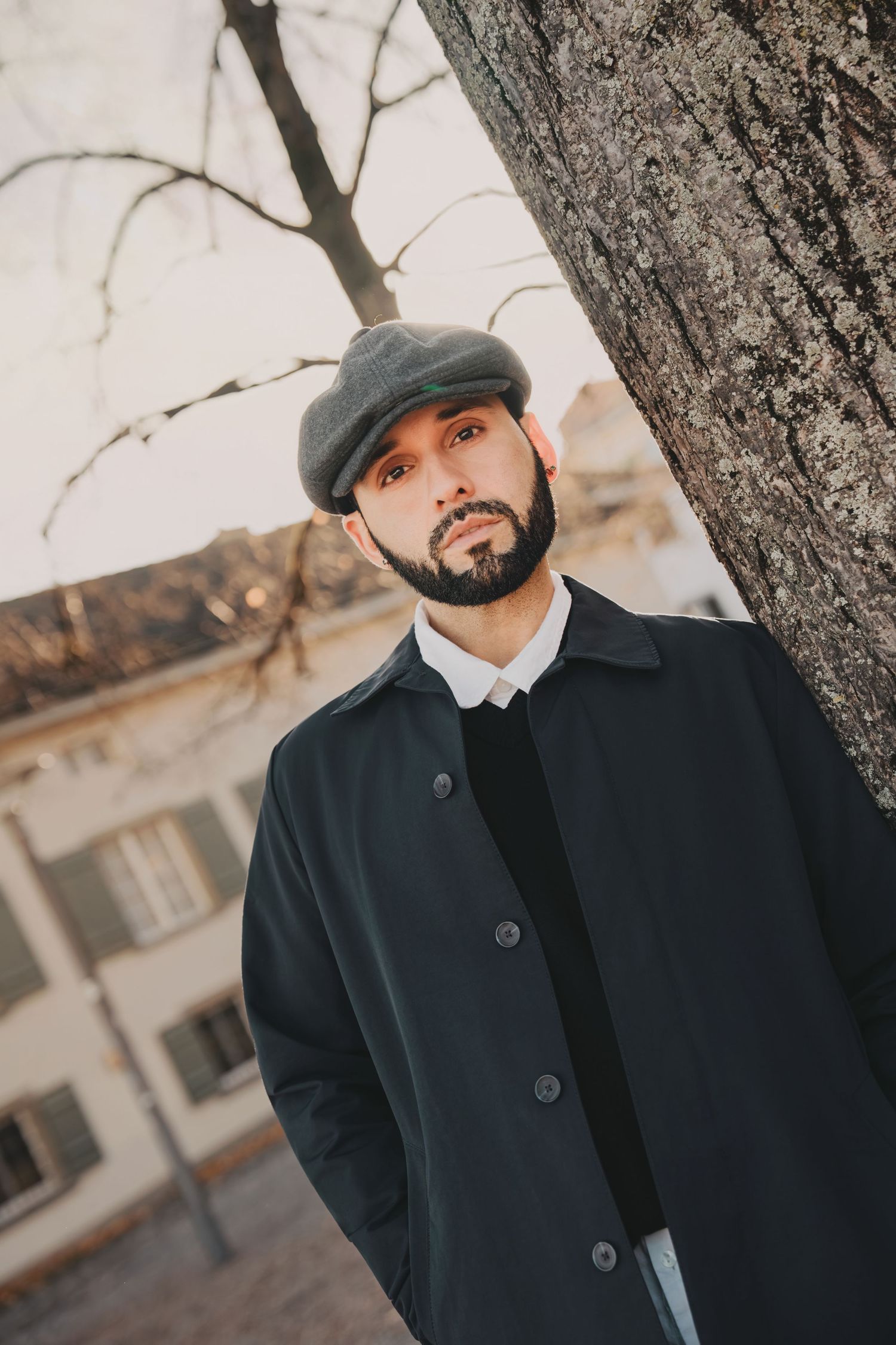 Bearded man in dark coat and grey flat cap leaning against a tree trunk in an urban outdoor setting.