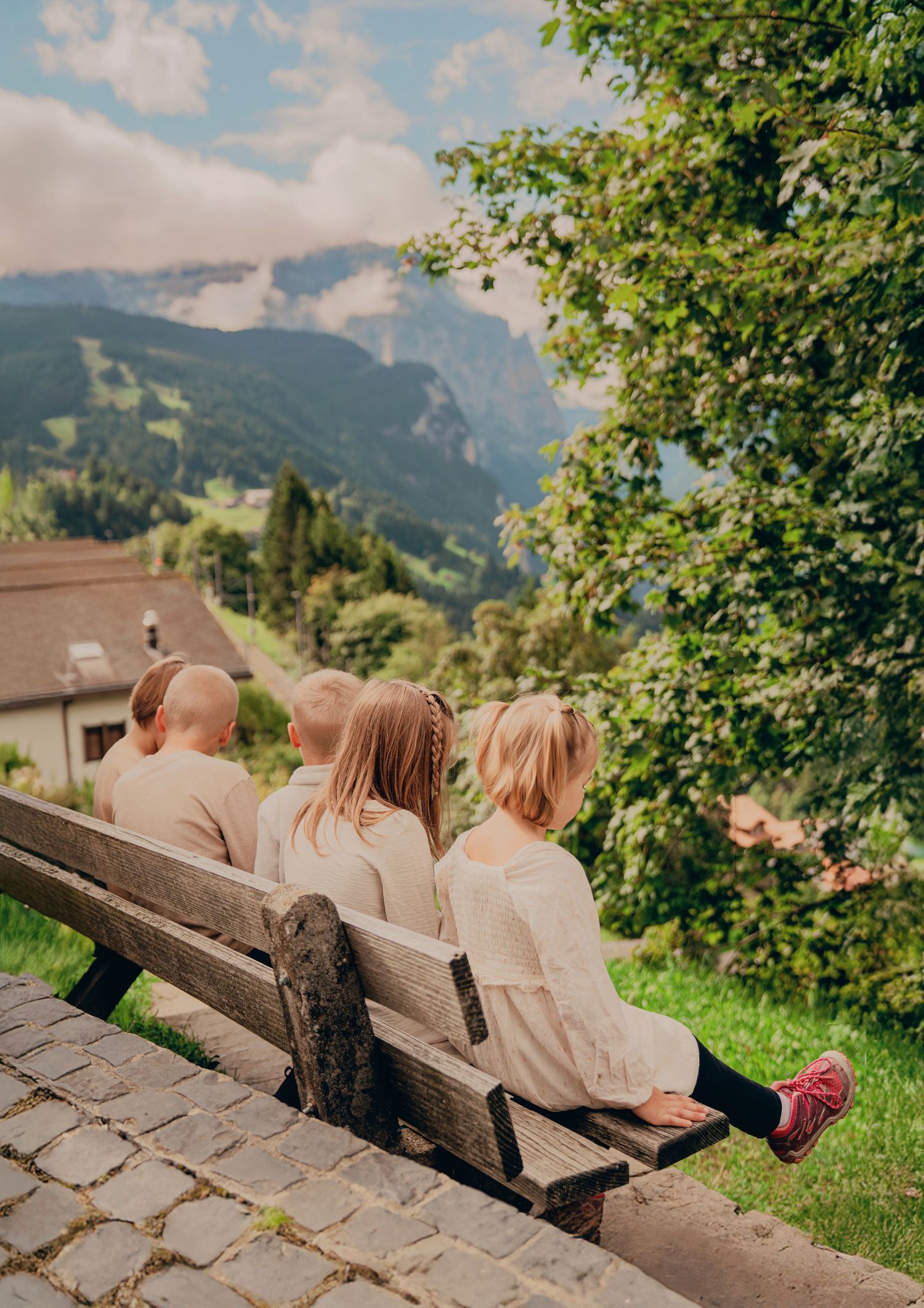 Four children sitting on a wooden bench admiring a lush Swiss mountain landscape.