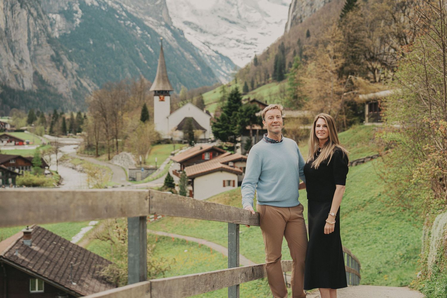 Couple standing together on a wooden bridge with a charming Swiss alpine village and snow-capped mountains behind them.