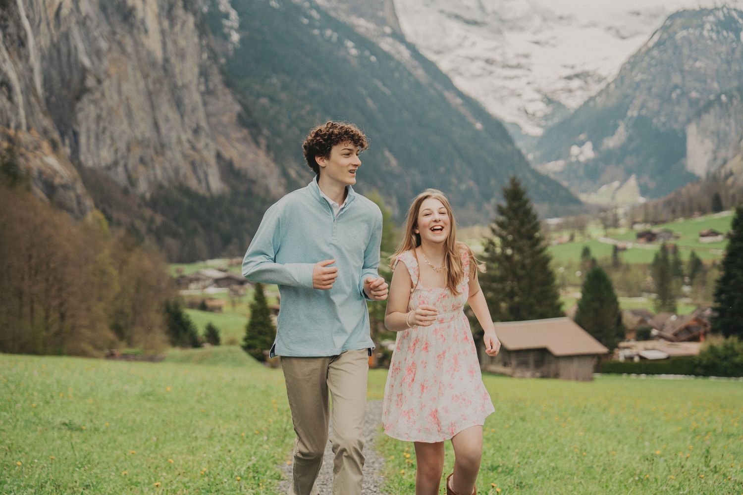Happy couple laughing and running through a green Alpine meadow with snow-capped mountains and Swiss village in background.