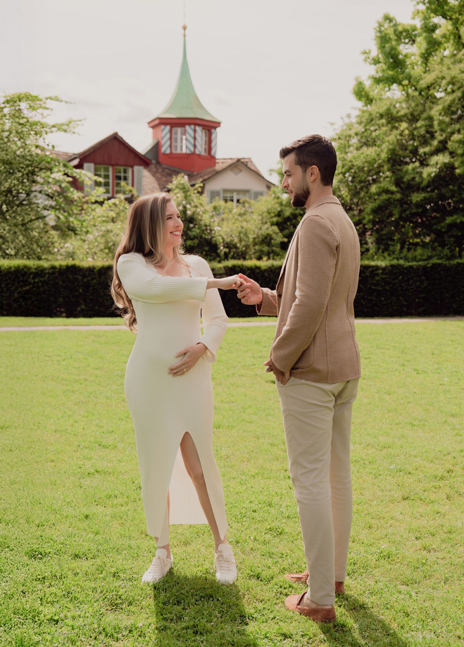 Expecting couple sharing a tender moment on green lawn with a red-towered building backdrop.