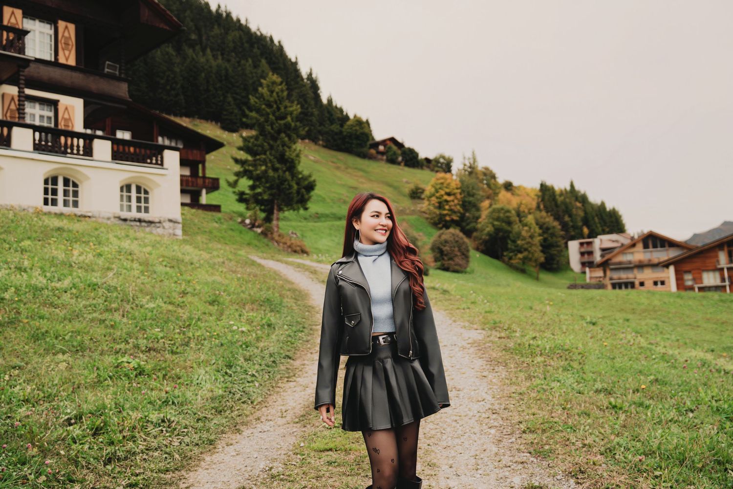 Young woman in black leather jacket and skirt strolling along a path in an alpine village setting.