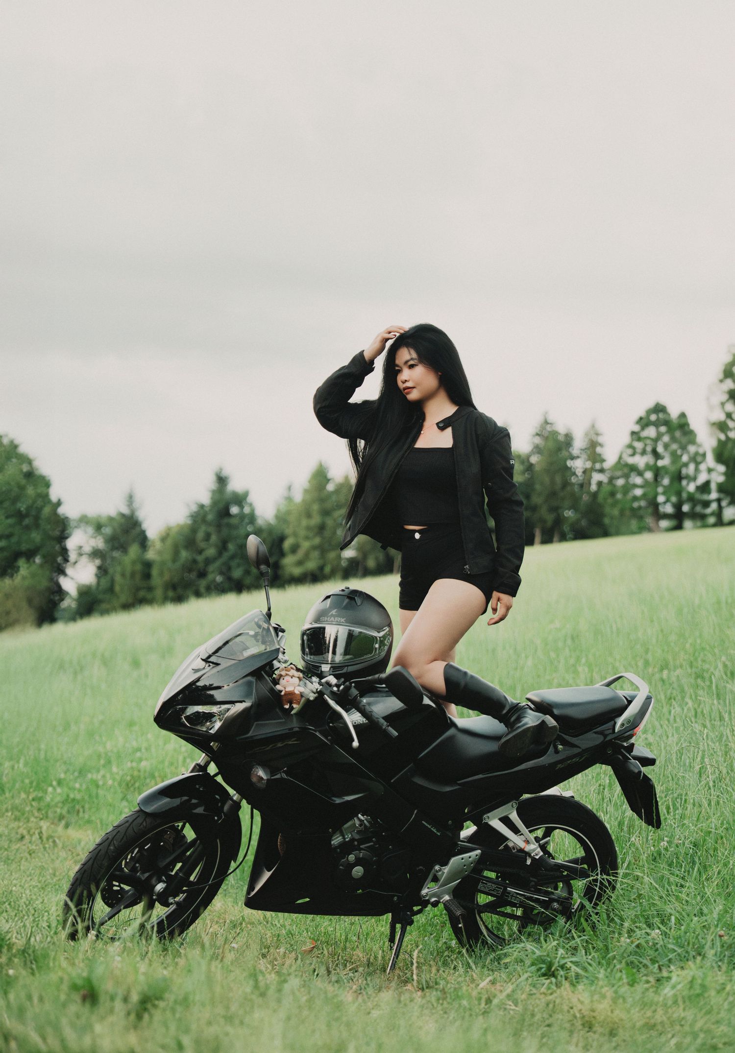 Stylish woman in black jacket and shorts posing with black sport motorcycle on green field.