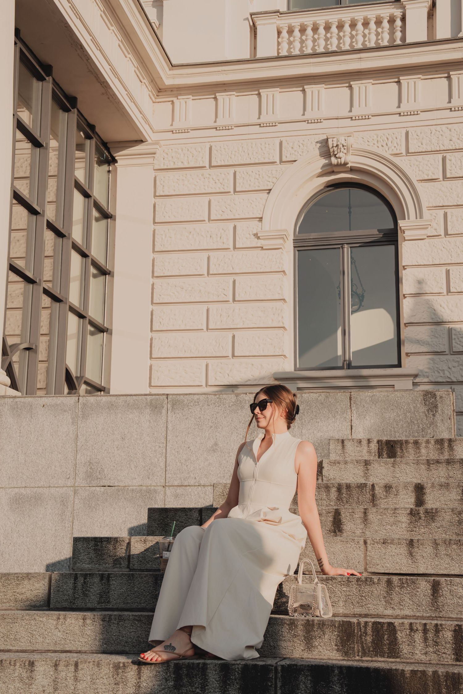 Woman in white dress and sunglasses sitting on steps of grand neoclassical building.