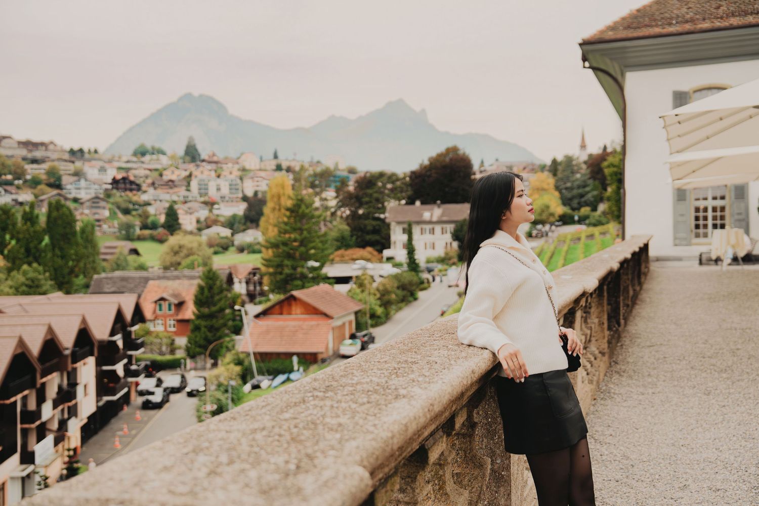 Elegant woman in cream top and black skirt gazes over charming Swiss mountain village from terrace.