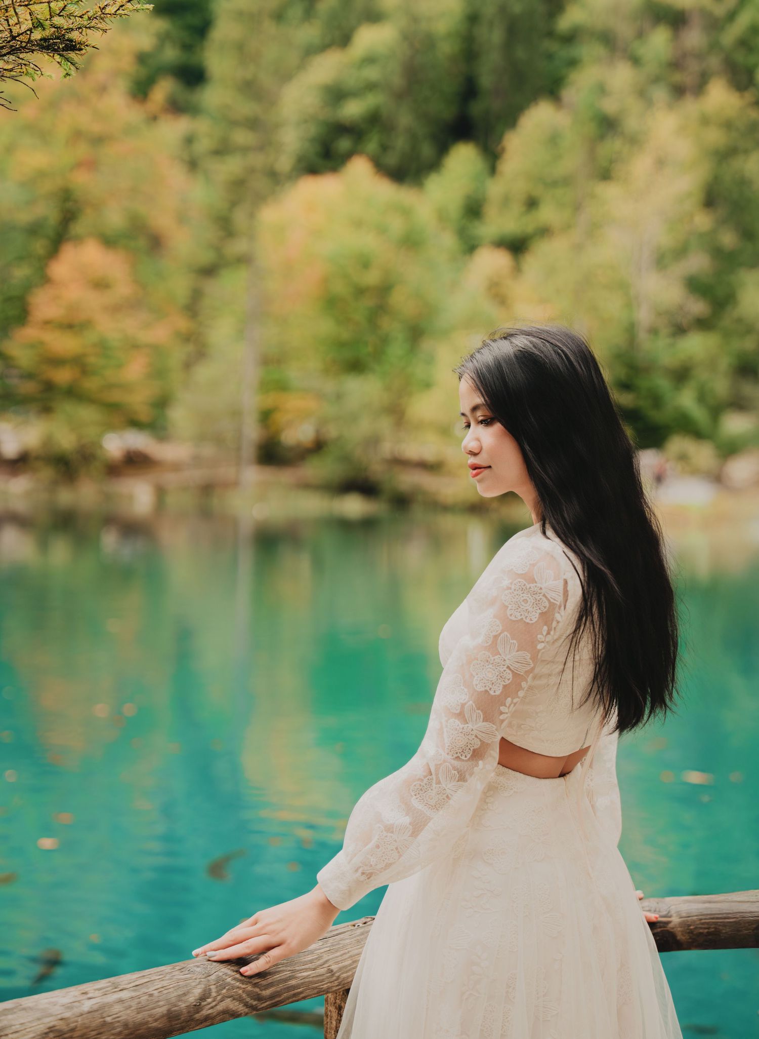 Woman in white lace dress gazes over turquoise lake surrounded by autumn forest.