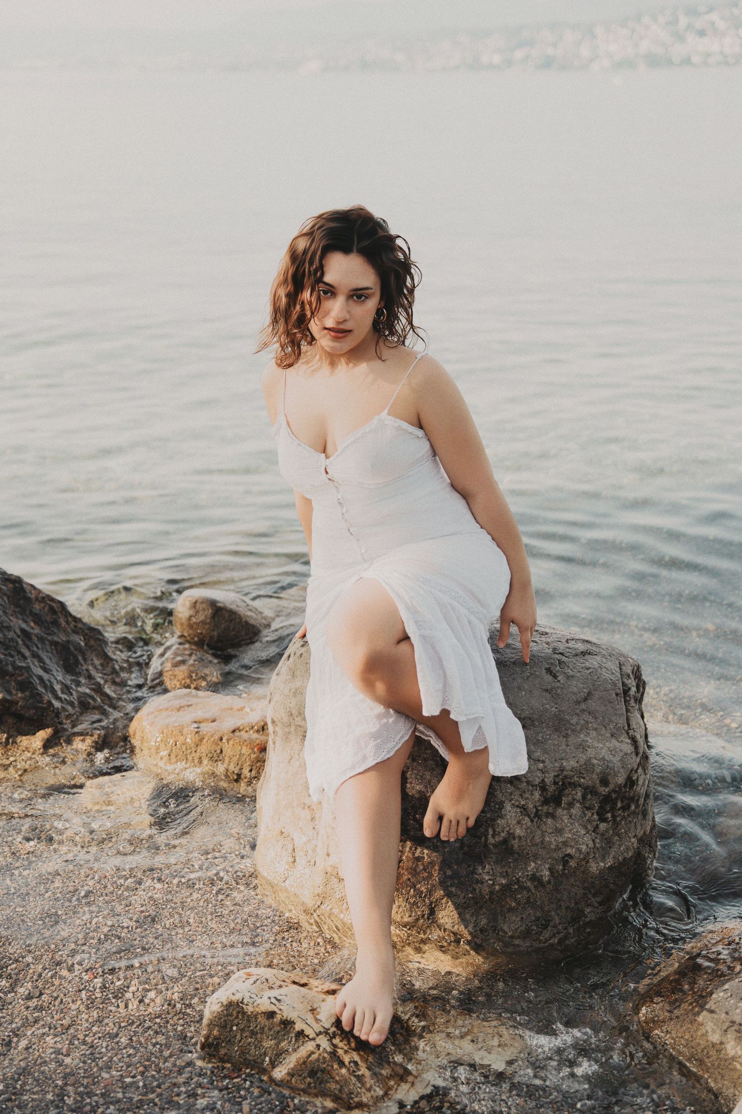 Brunette woman in white dress sitting on seaside rocks, gazing at camera.
