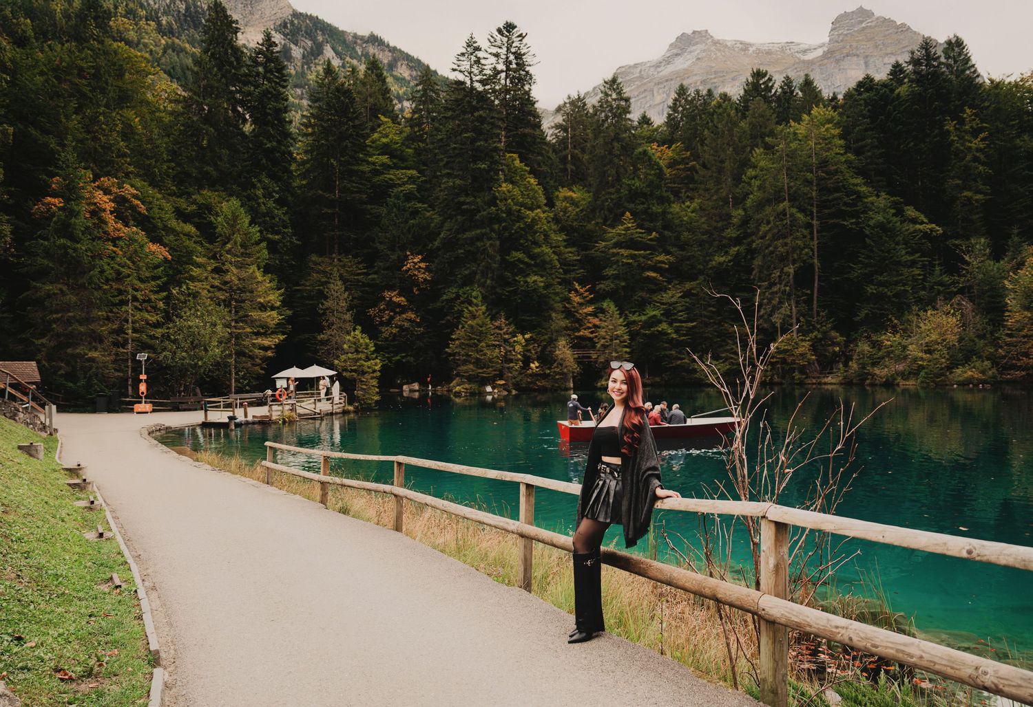 Woman in black outfit poses by turquoise alpine lake with wooden fence, mountains and forest backdrop.