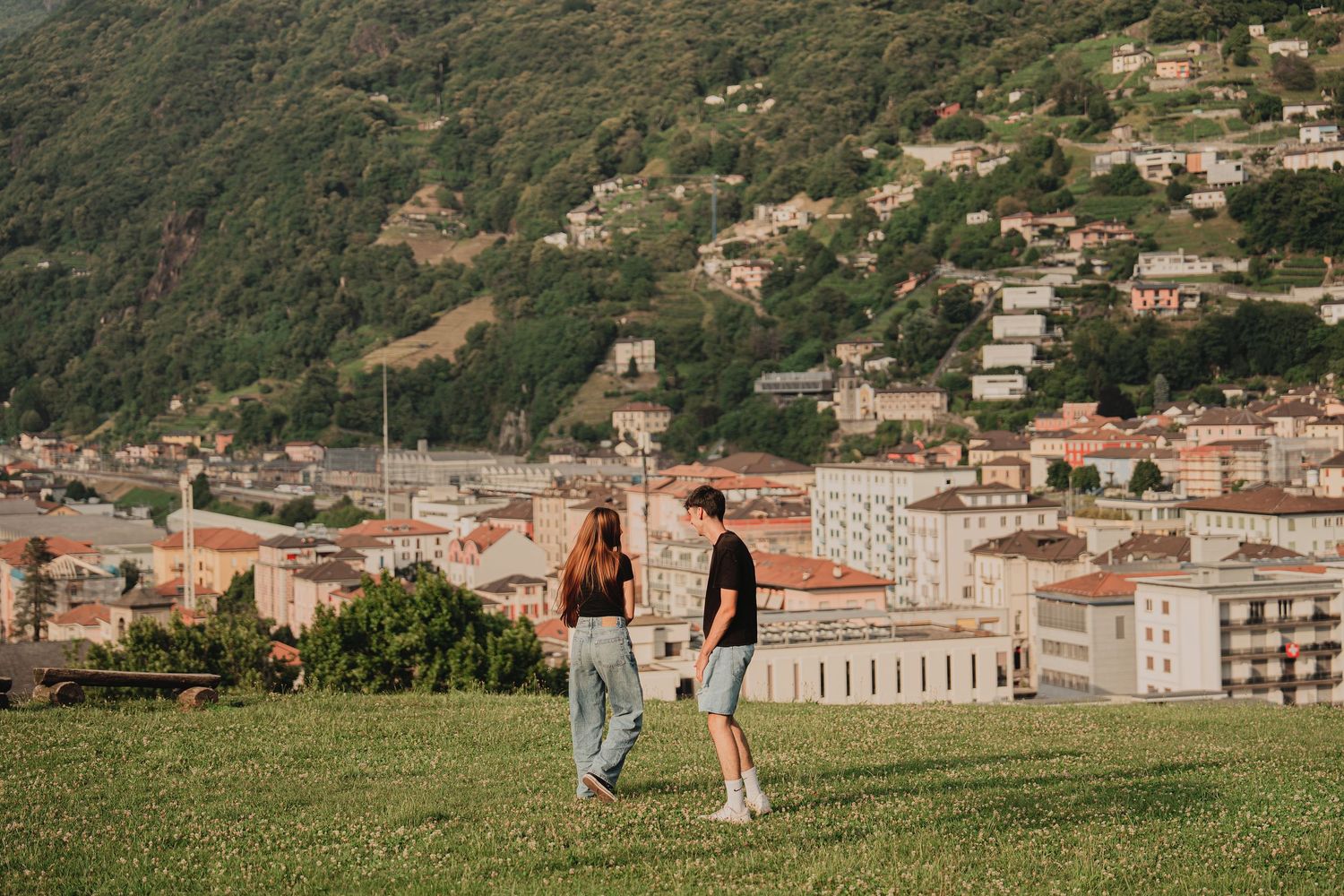 Young couple strolling across a green meadow with a scenic European hillside village in the background.