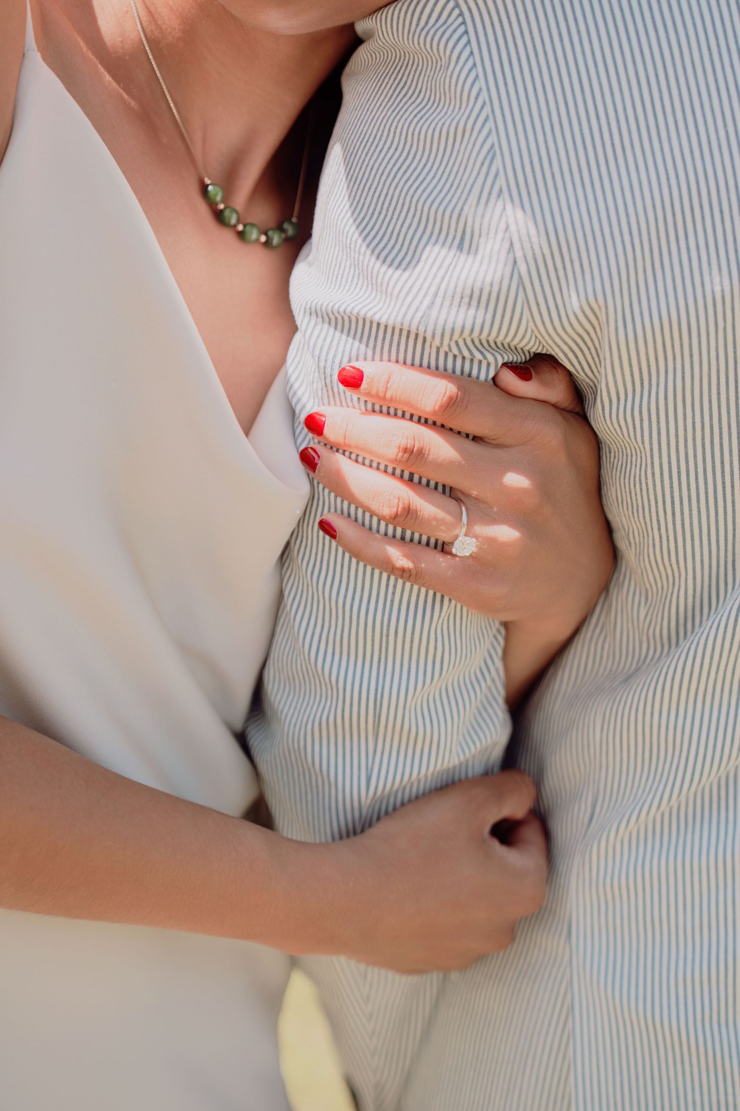 Close-up of bride embracing groom, showcasing diamond engagement ring, red nails, and delicate necklace.