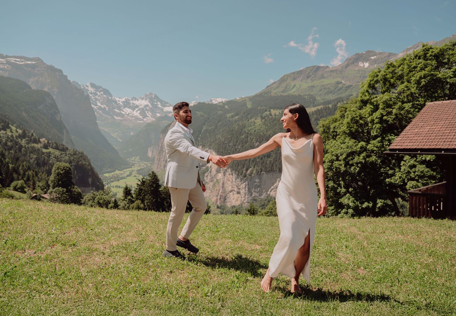 Elegant couple holding hands and walking on a sunlit alpine meadow with majestic mountain valley views.