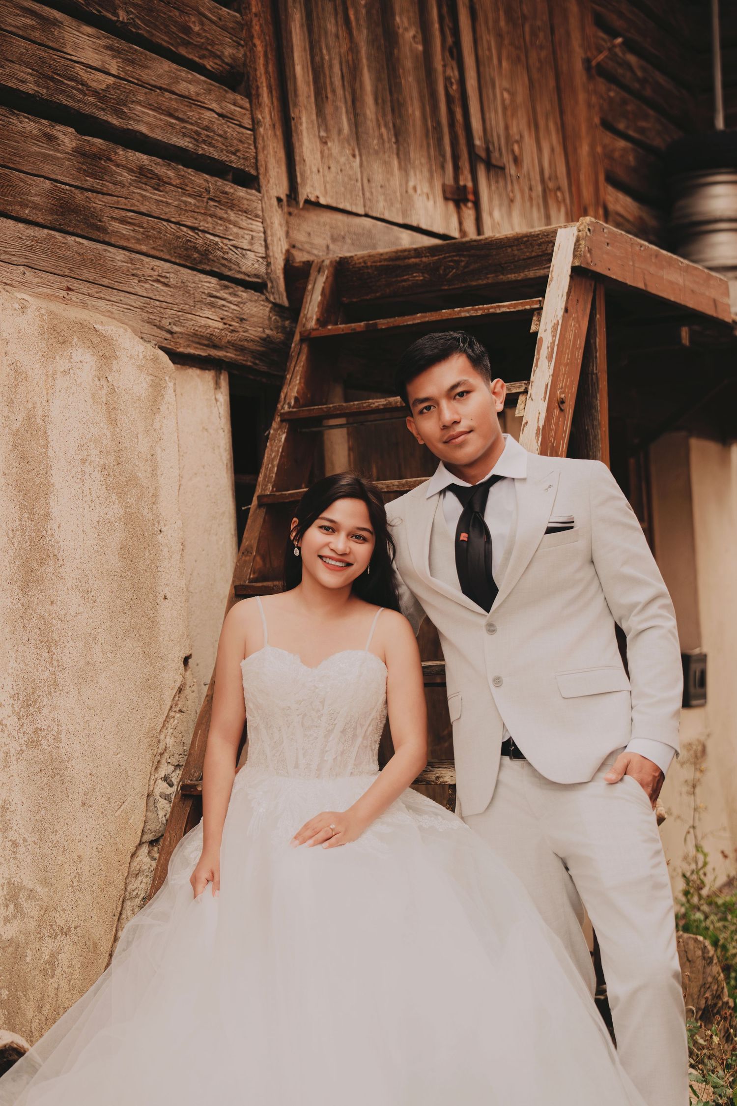 Smiling bride in strapless white gown and groom in light suit pose by rustic wooden steps.