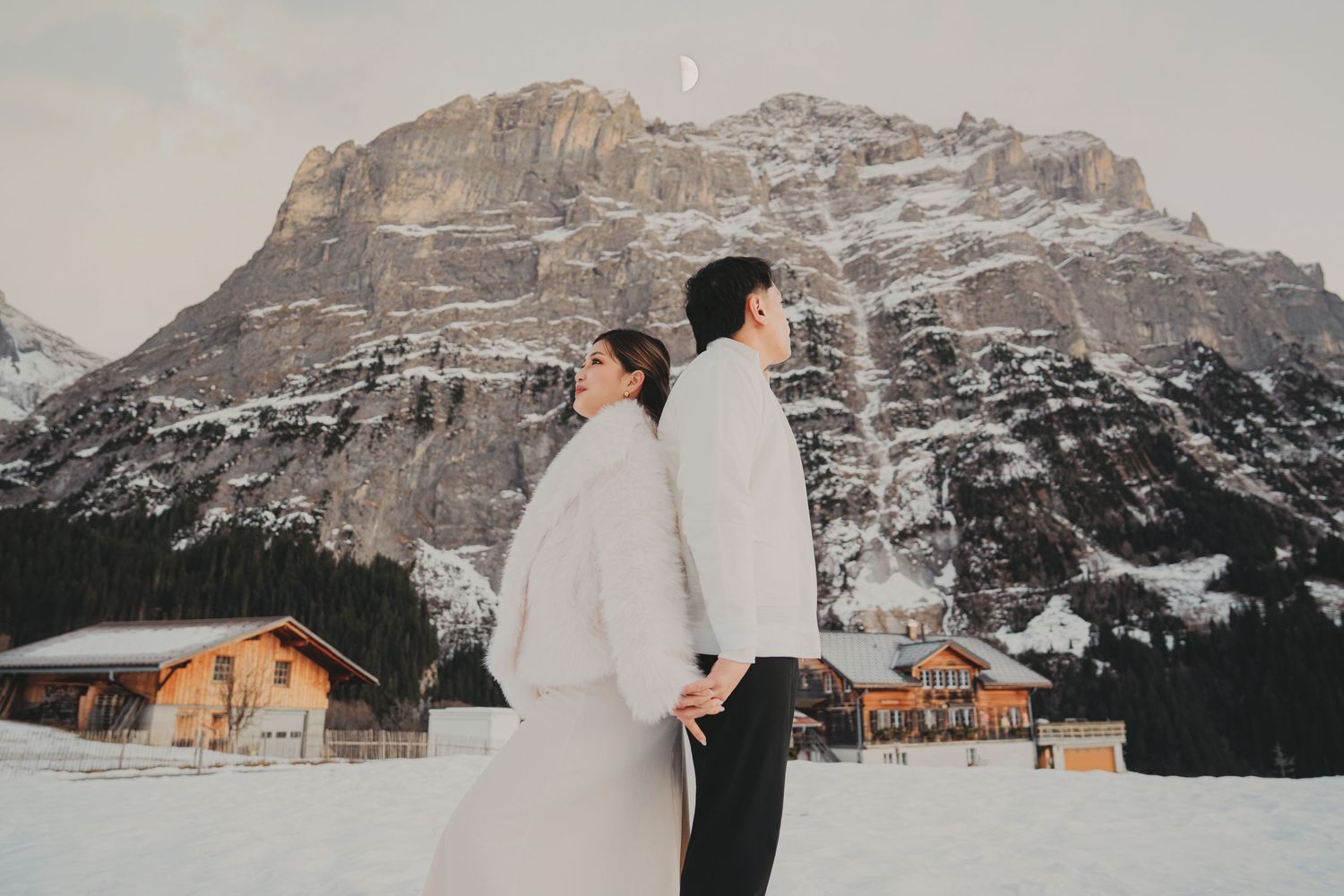 Couple standing back-to-back holding hands in winter attire with snow-covered Alpine mountain backdrop.