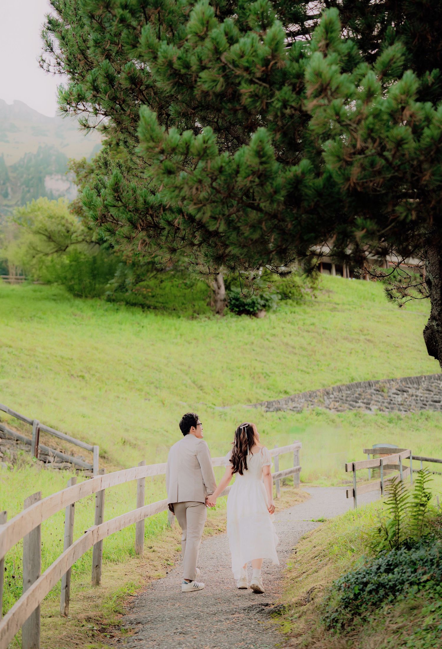 Couple in wedding attire holding hands walking down a path surrounded by lush green hills and a pine tree.