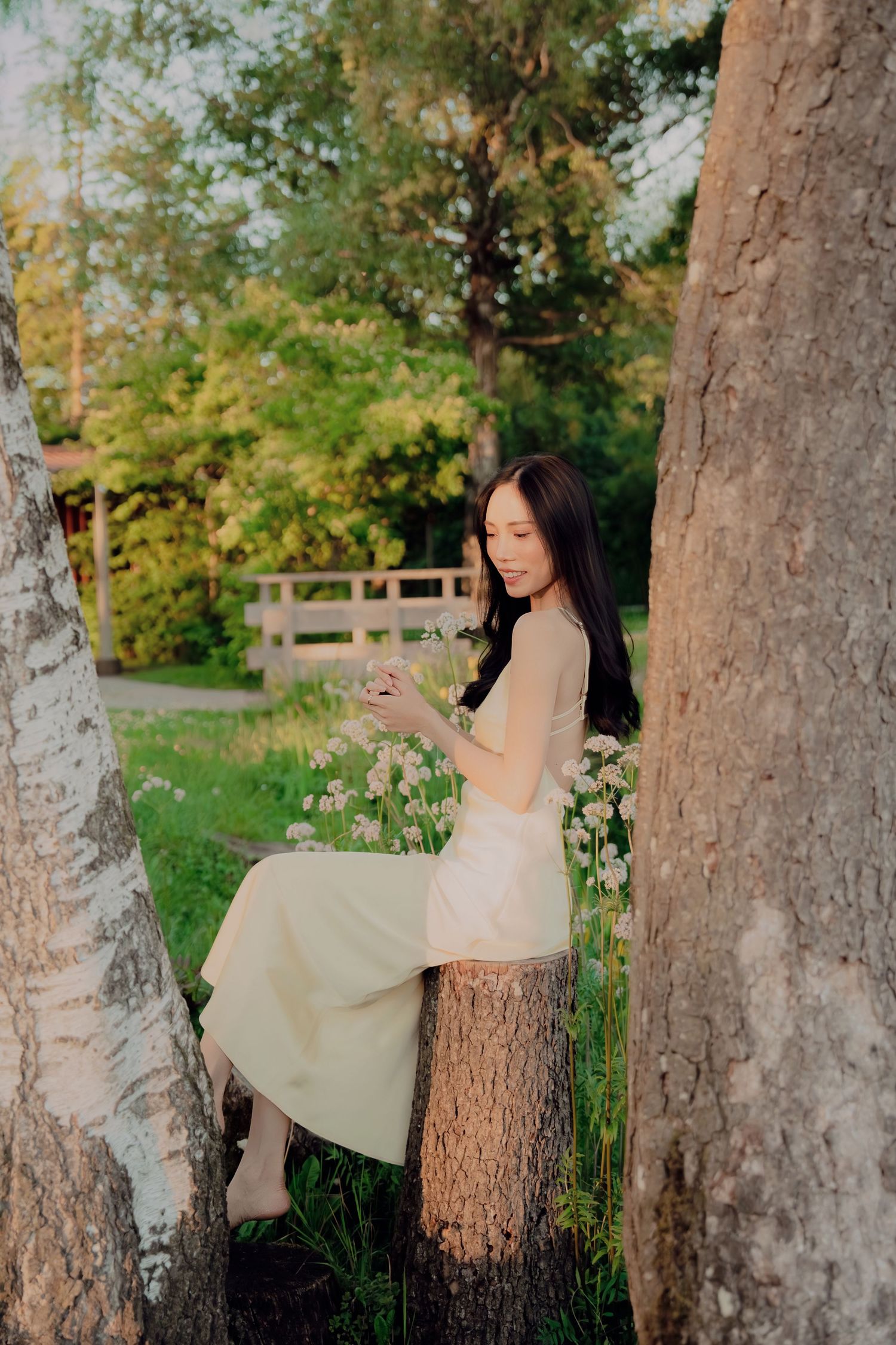 Young woman in pale yellow dress sits on a tree stump holding flowers, surrounded by lush greenery.