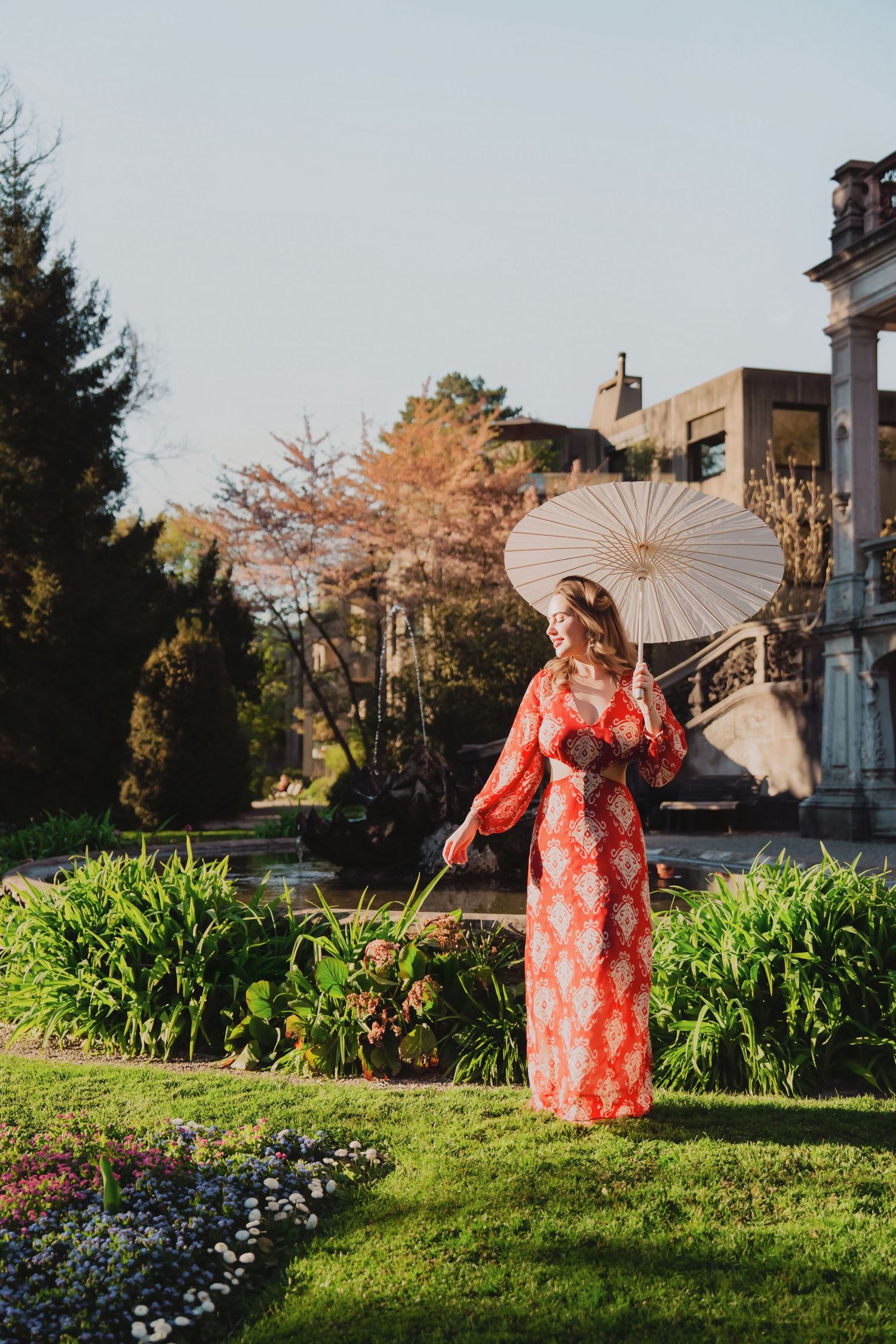 Woman in red floral kimono holding white parasol in lush garden with historic building backdrop.