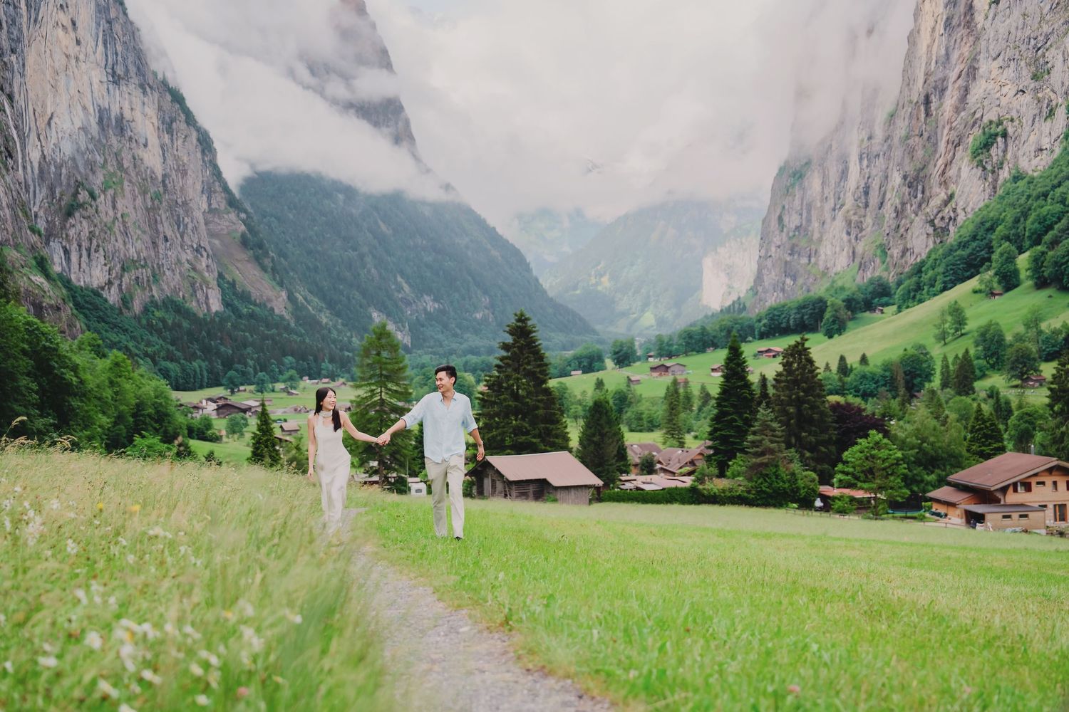 Happy couple walking hand-in-hand through scenic Lauterbrunnen valley with misty Swiss Alps and green fields.
