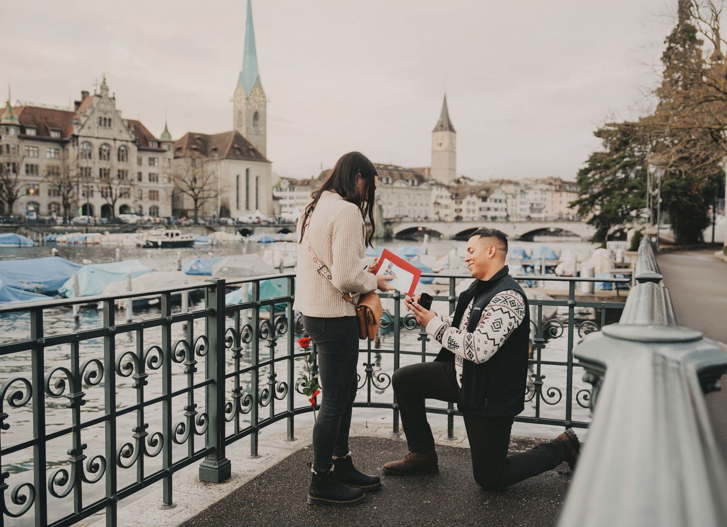 Man proposing on one knee to woman on bridge in Zurich with historic cityscape backdrop.