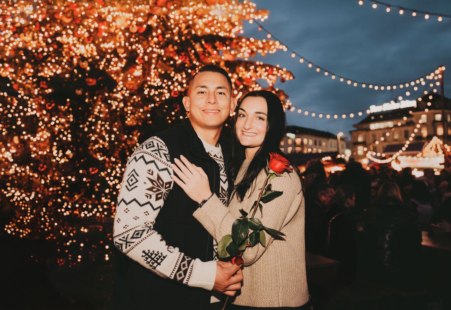 Couple smiling together holding a red rose near a glowing Christmas tree at night.