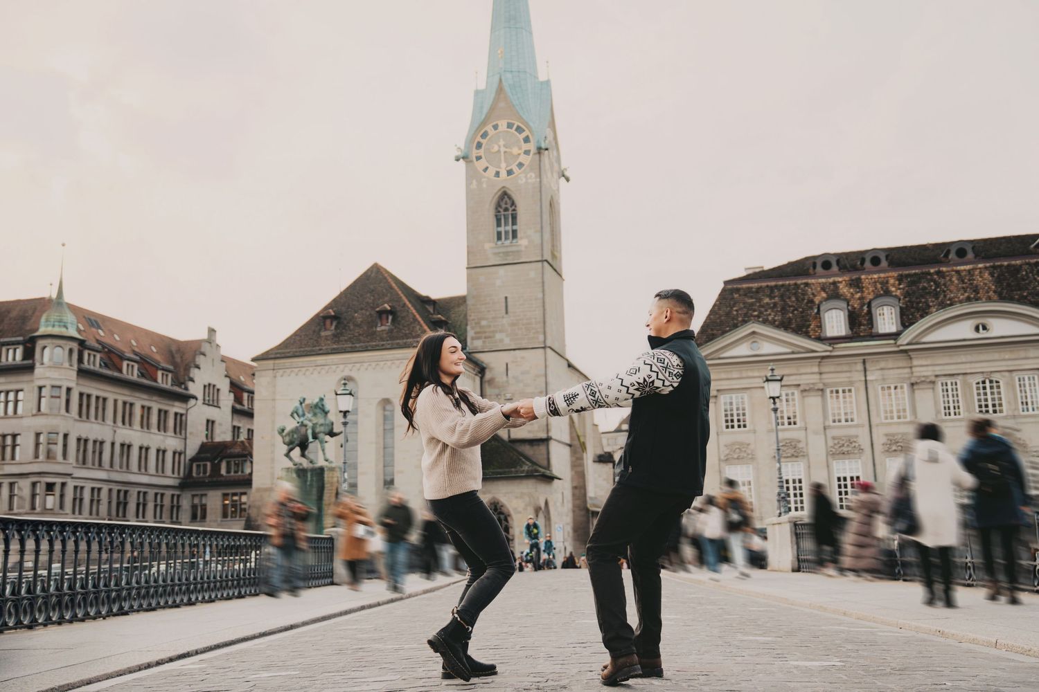 Playful couple spinning on a cobblestone bridge in Zurich with church tower backdrop.