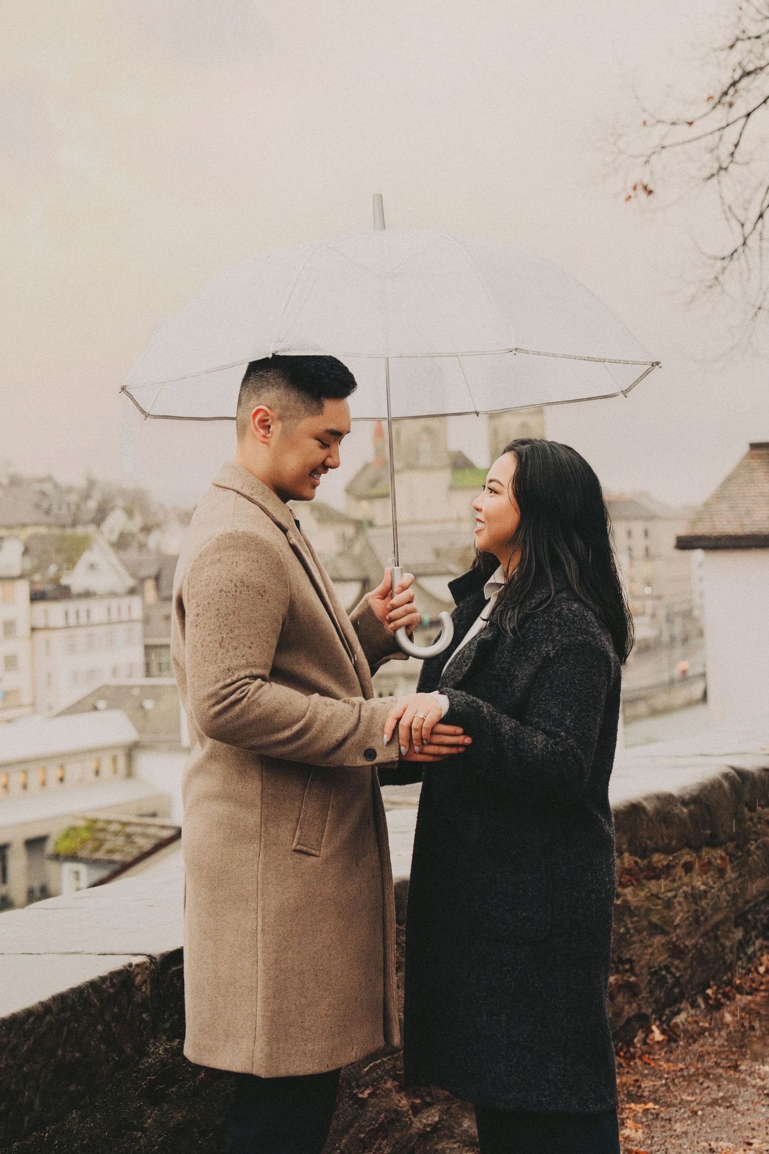 Couple holding hands and sharing a tender moment under a clear umbrella in rainy Zurich.