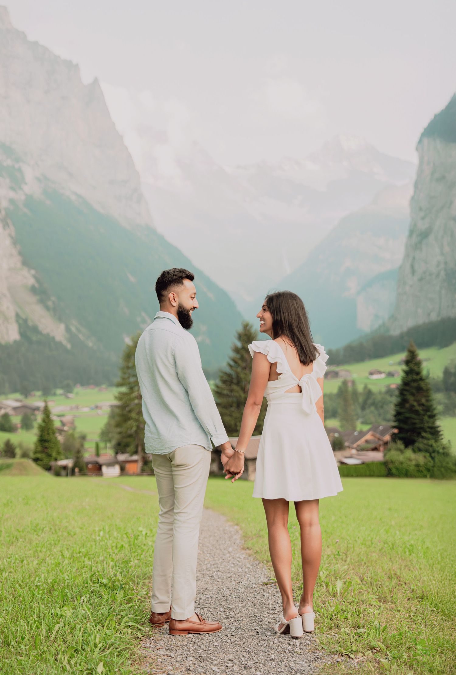Couple holding hands on scenic path facing misty Swiss Alps mountains with green valley and pine trees in background.