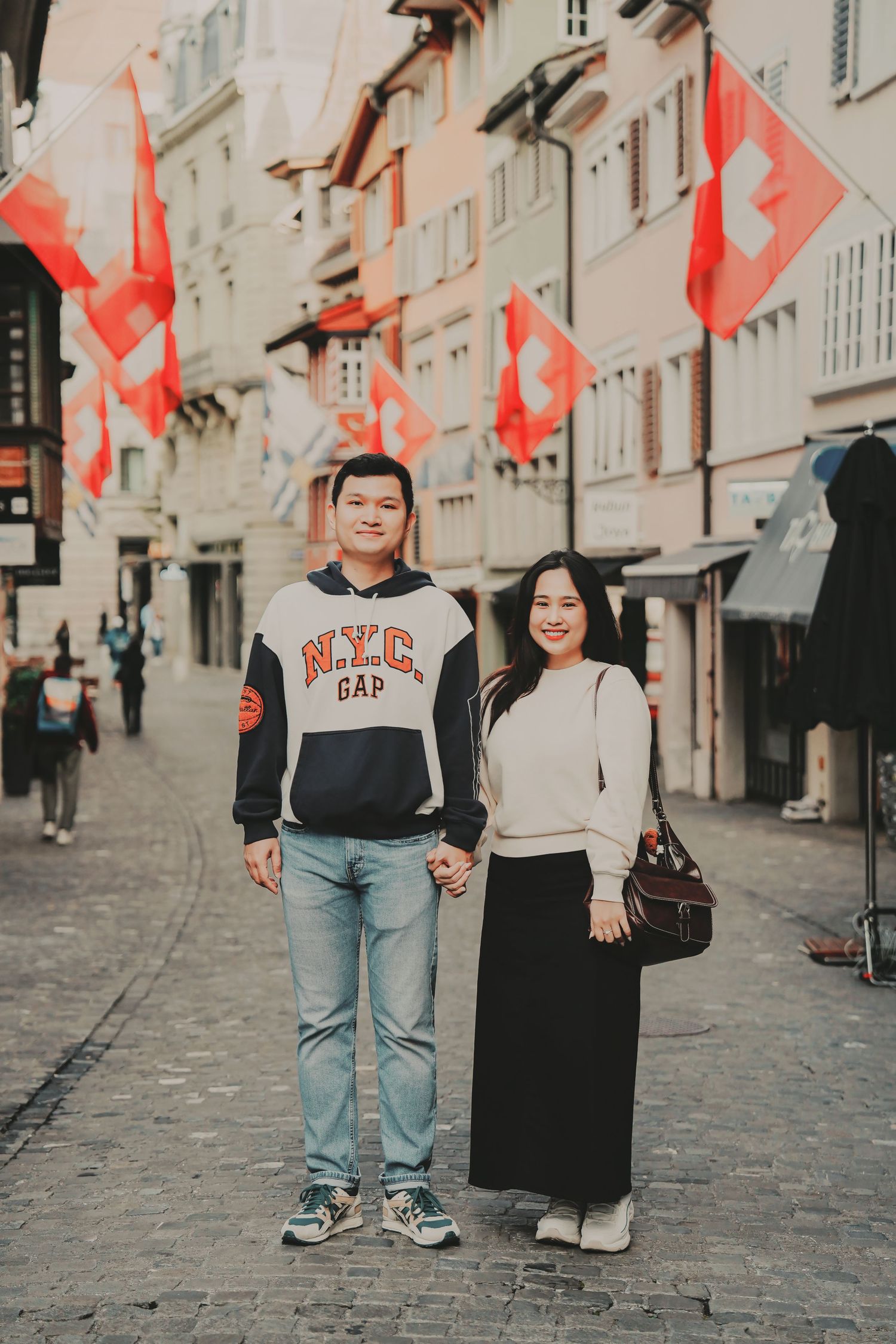 A smiling couple poses hand-in-hand on a Zurich cobblestone street decorated with Swiss flags.
