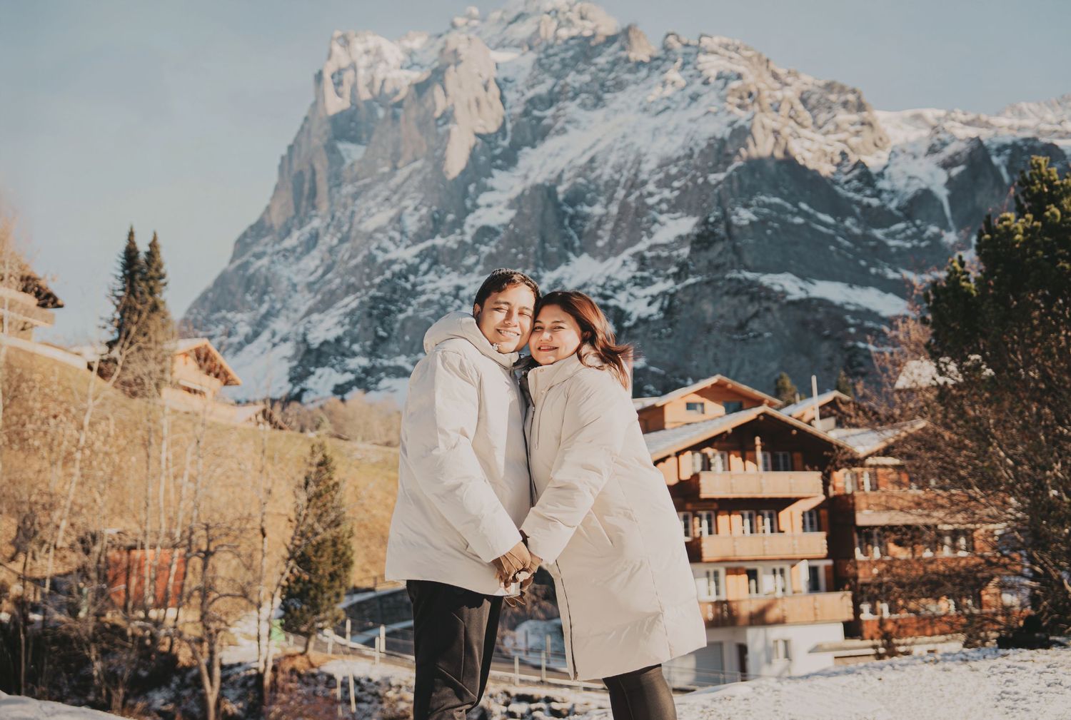 Couple embracing and smiling in snowy Swiss mountain village wearing matching white coats.