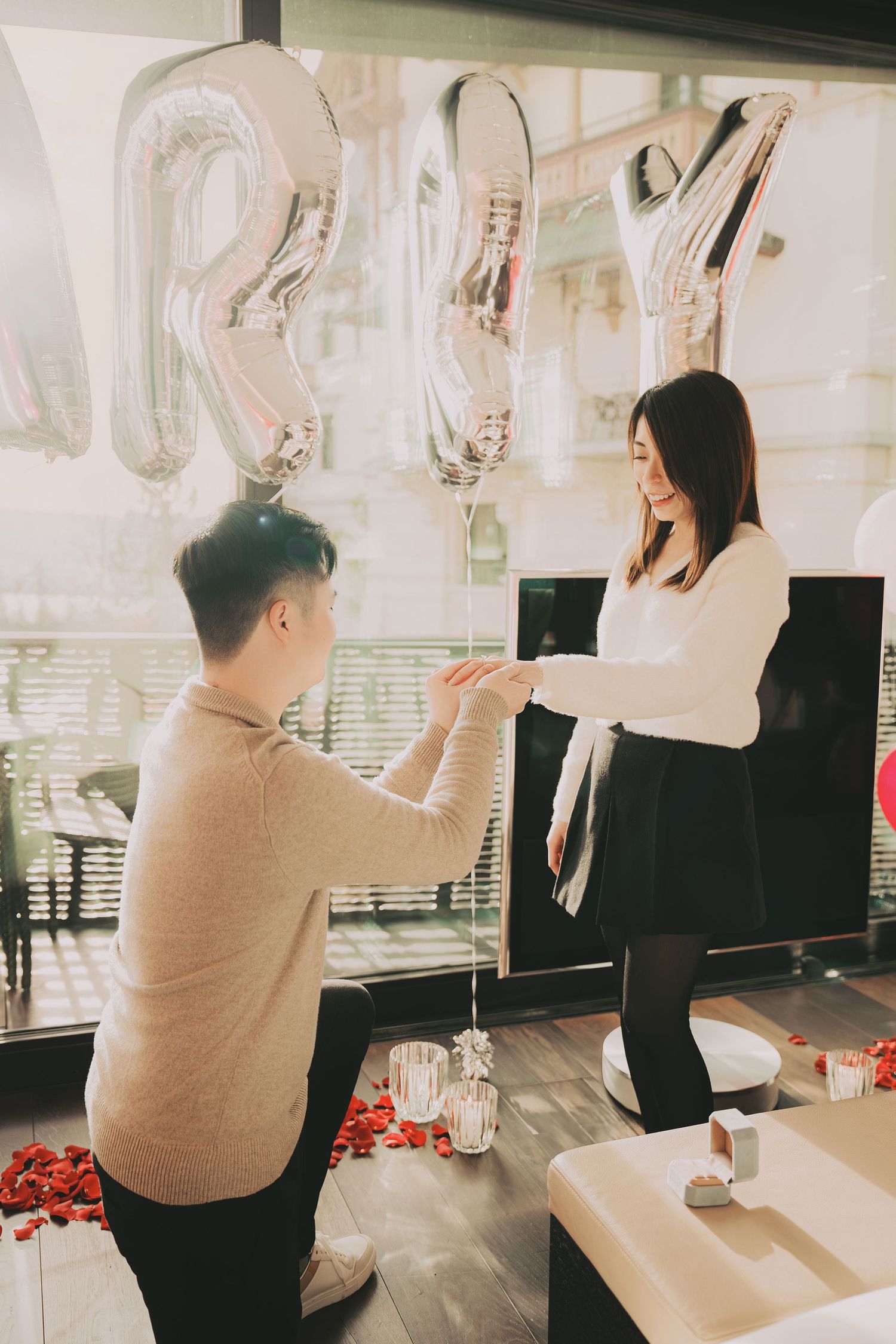 Man kneeling on one knee proposing to his girlfriend in a romantic room decorated with MARRY balloons and rose petals.