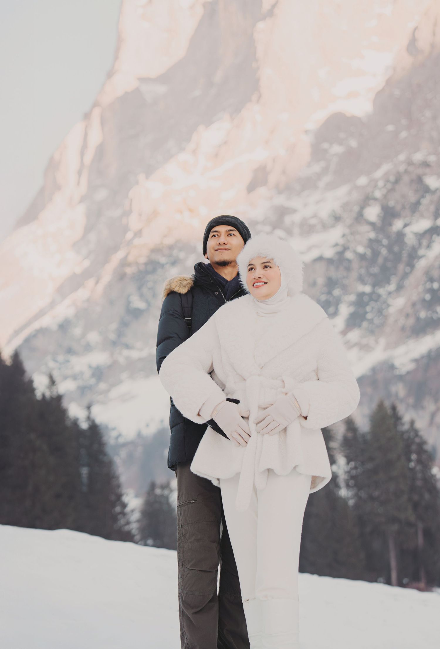 Couple posing in snowy mountain setting, woman in white outfit and man in dark jacket embracing warmly.