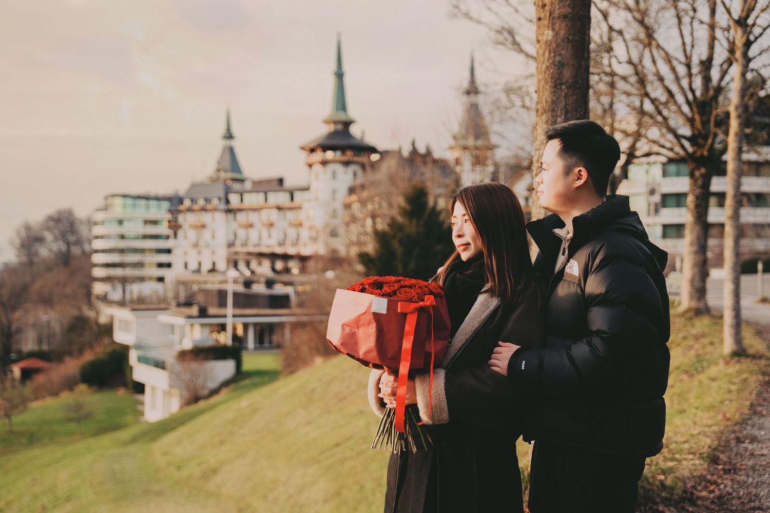 Couple embracing while holding red gift box on hillside with castle hotel backdrop.