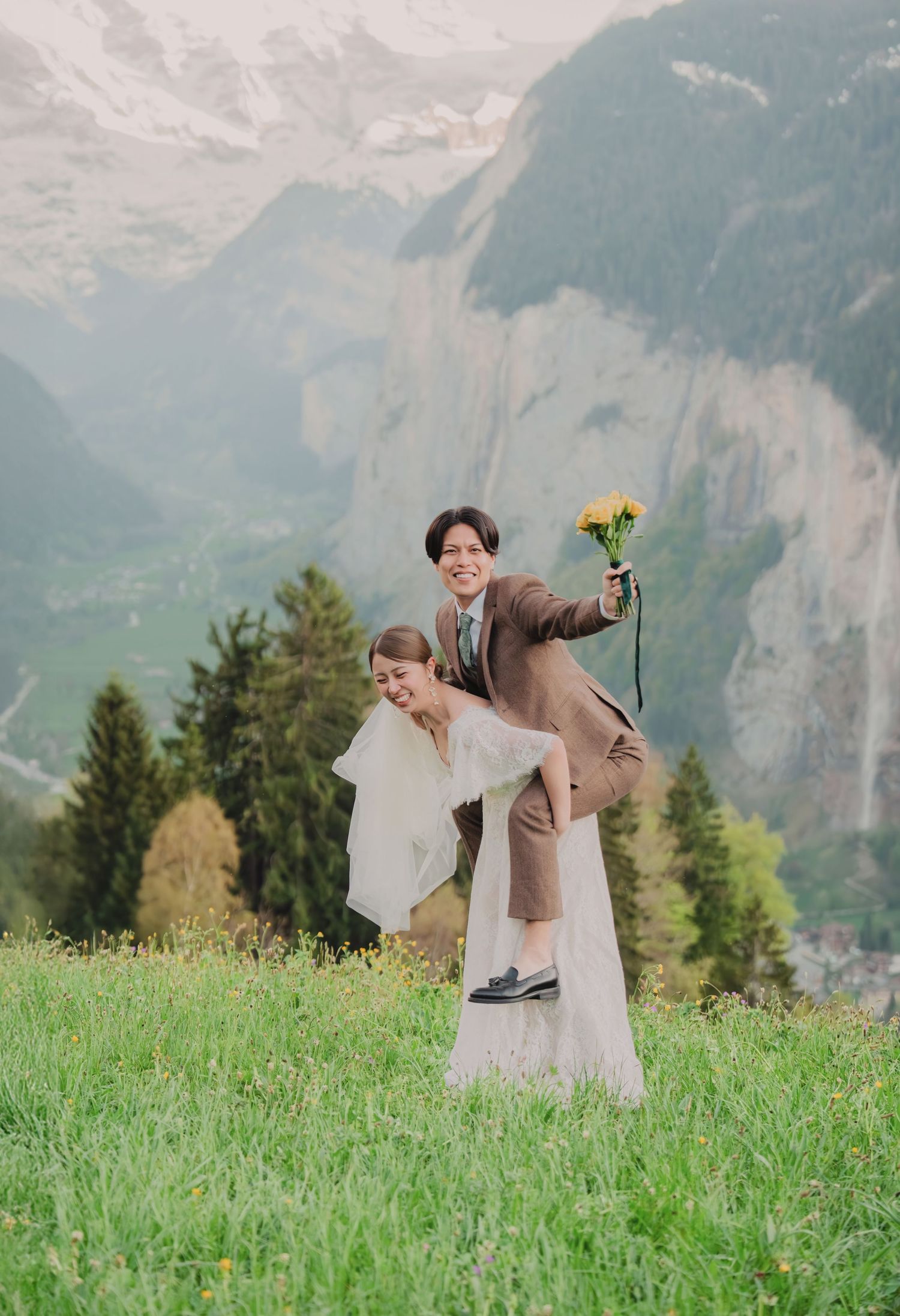 Joyful couple in wedding attire posing on a green alpine meadow with misty Swiss mountain cliffs in the background.